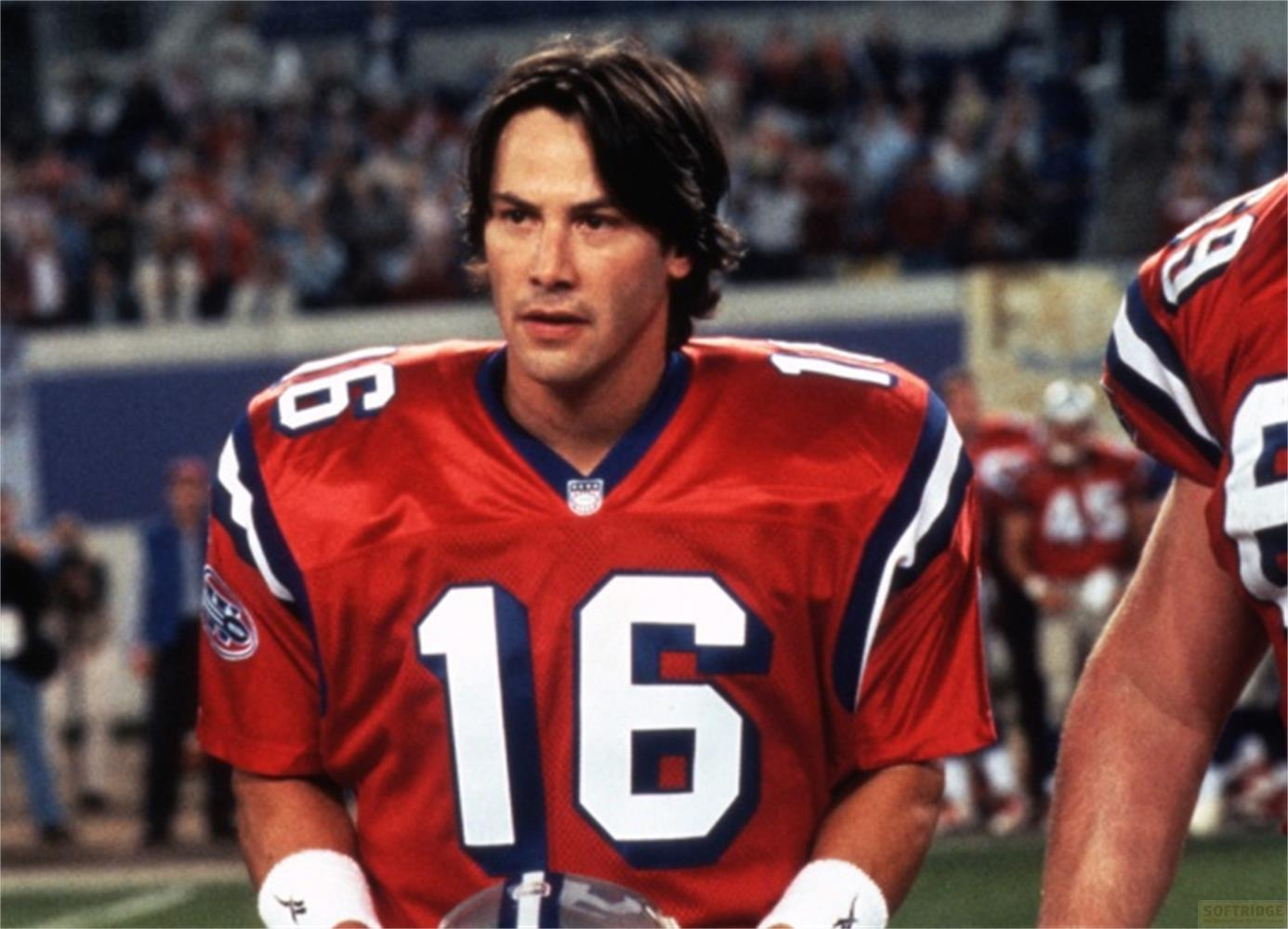 Keanu Reeves, con una maglia da football americano rossa con il numero 16, in piedi su un campo con spettatori sfocati sullo sfondo.