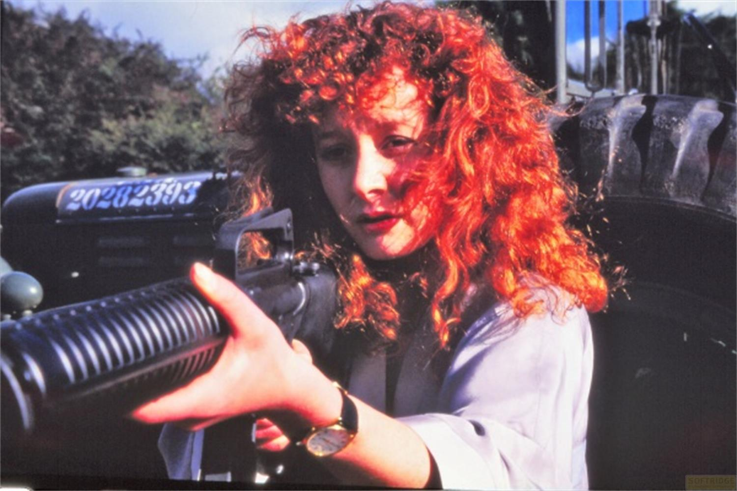 Donna con capelli ricci rossi tiene una pistola, pneumatico di trattore e targa sullo sfondo.