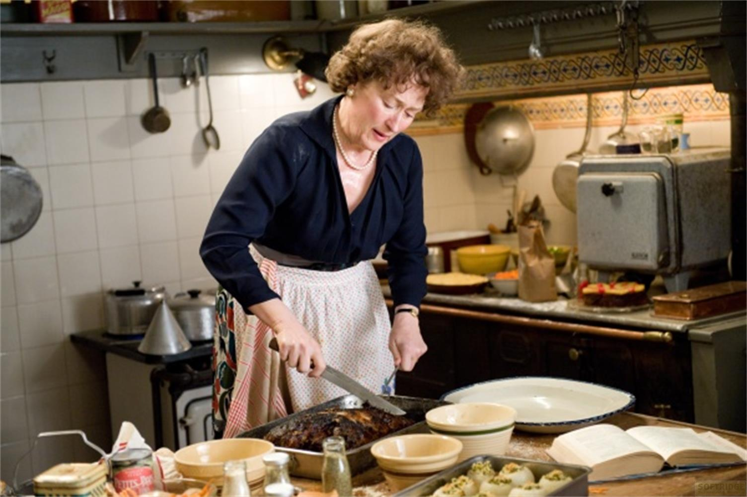 Una donna con capelli ricci castani, che indossa un top blu scuro e un grembiule bianco, taglia un piatto arrosto in una cucina rustica.