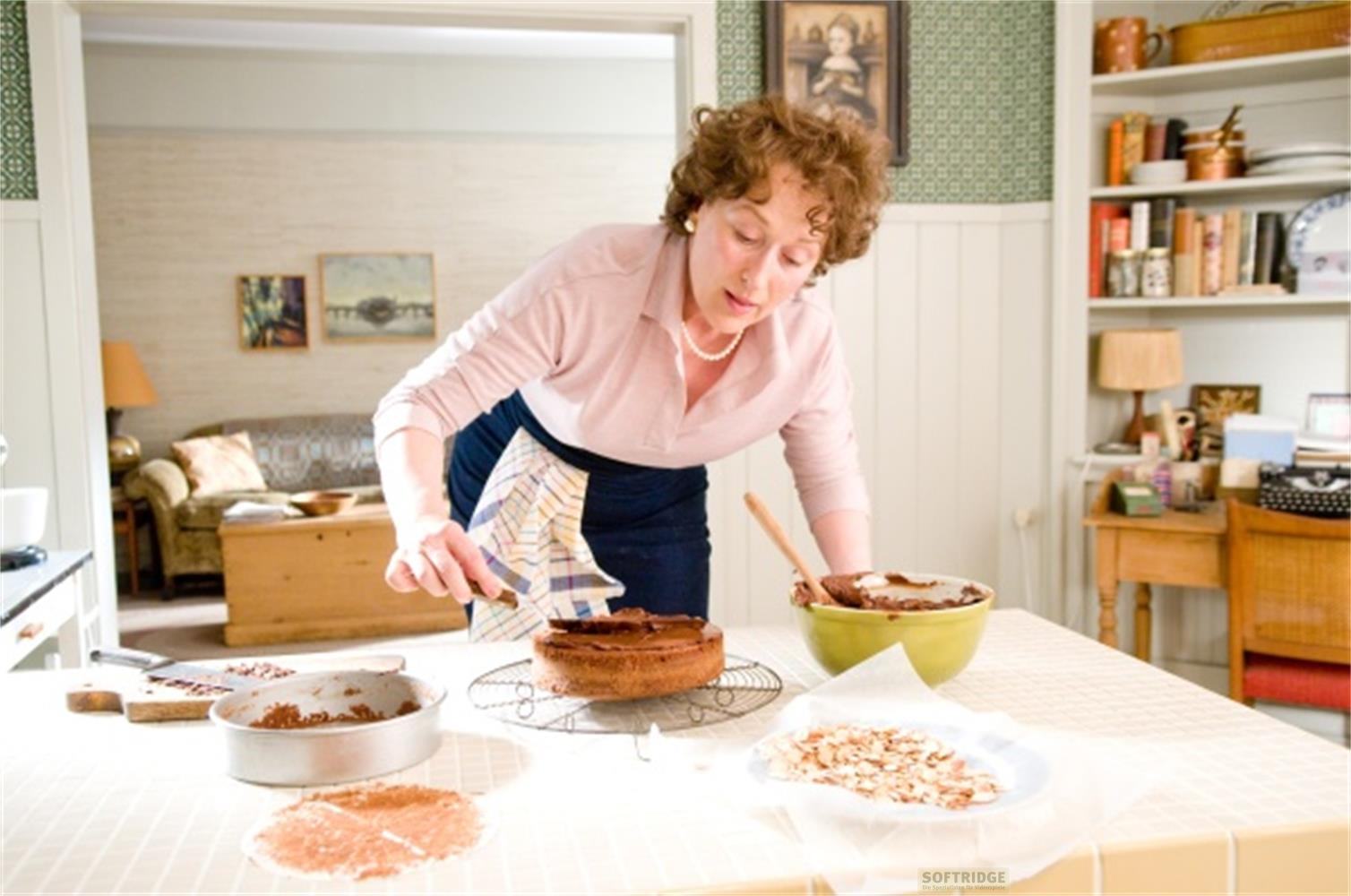 Una donna con capelli ricci, camicia rosa e collana di perle, decora una torta al cioccolato in cucina.