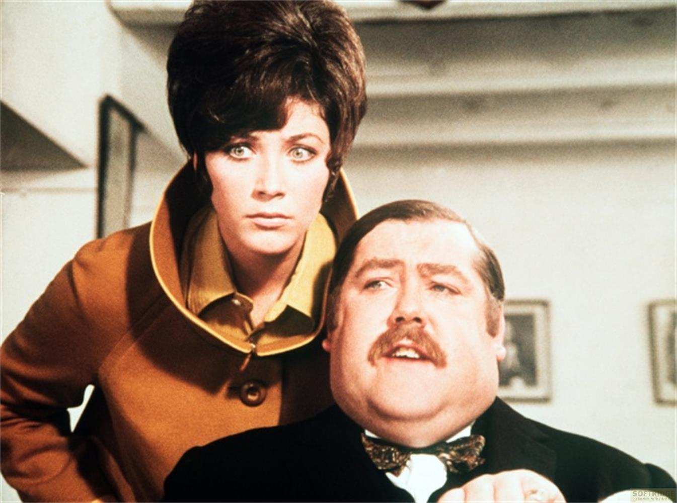 Un uomo con i baffi indossa un abito, una donna con i capelli castani indossa un cappotto marrone.