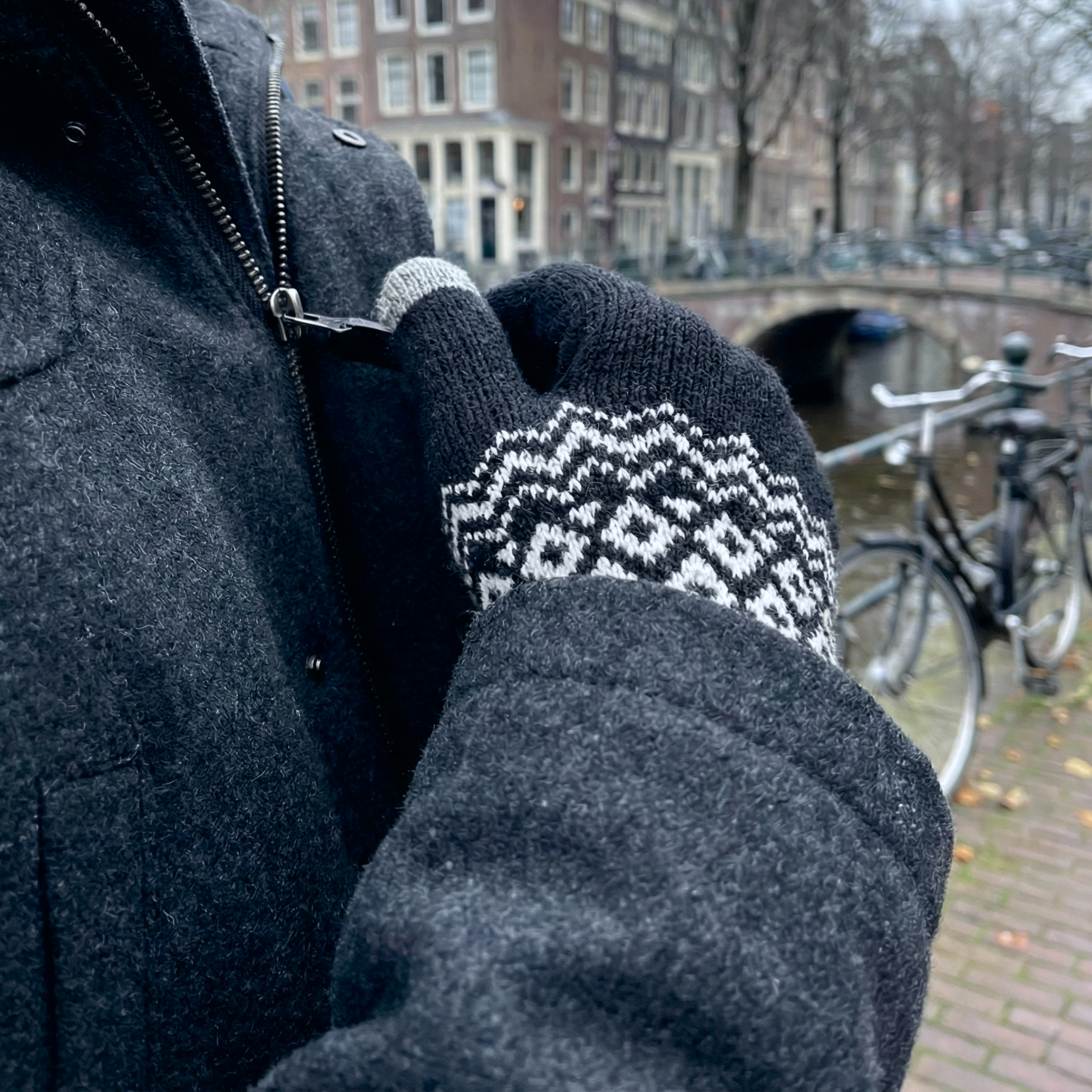 Person in dark coat zipping up, wearing patterned knitted gloves. City canal background.