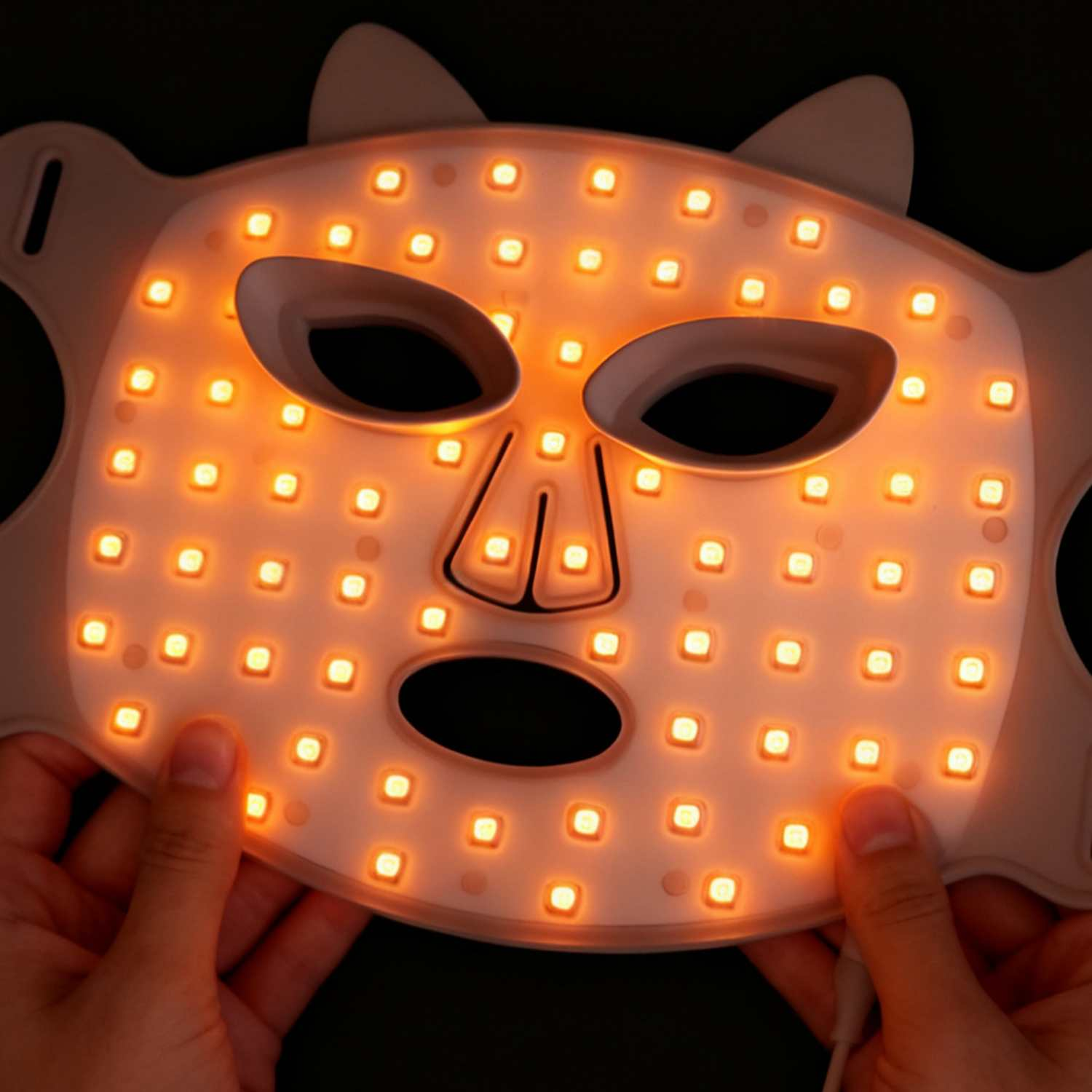 Hände halten eine weiße LED-Gesichtsmaske mit orangen Lichtern vor dunklem Hintergrund.