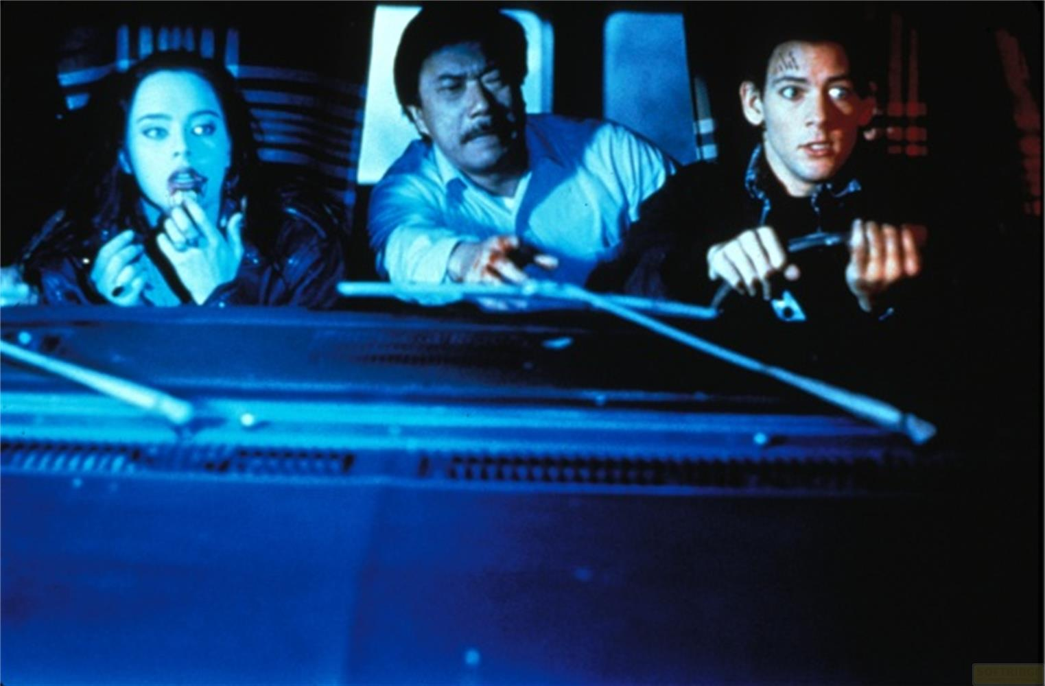 Tre persone in un'auto. La donna a sinistra guarda in basso. L'uomo al centro indossa una camicia azzurra. L'uomo a destra tiene uno strumento.