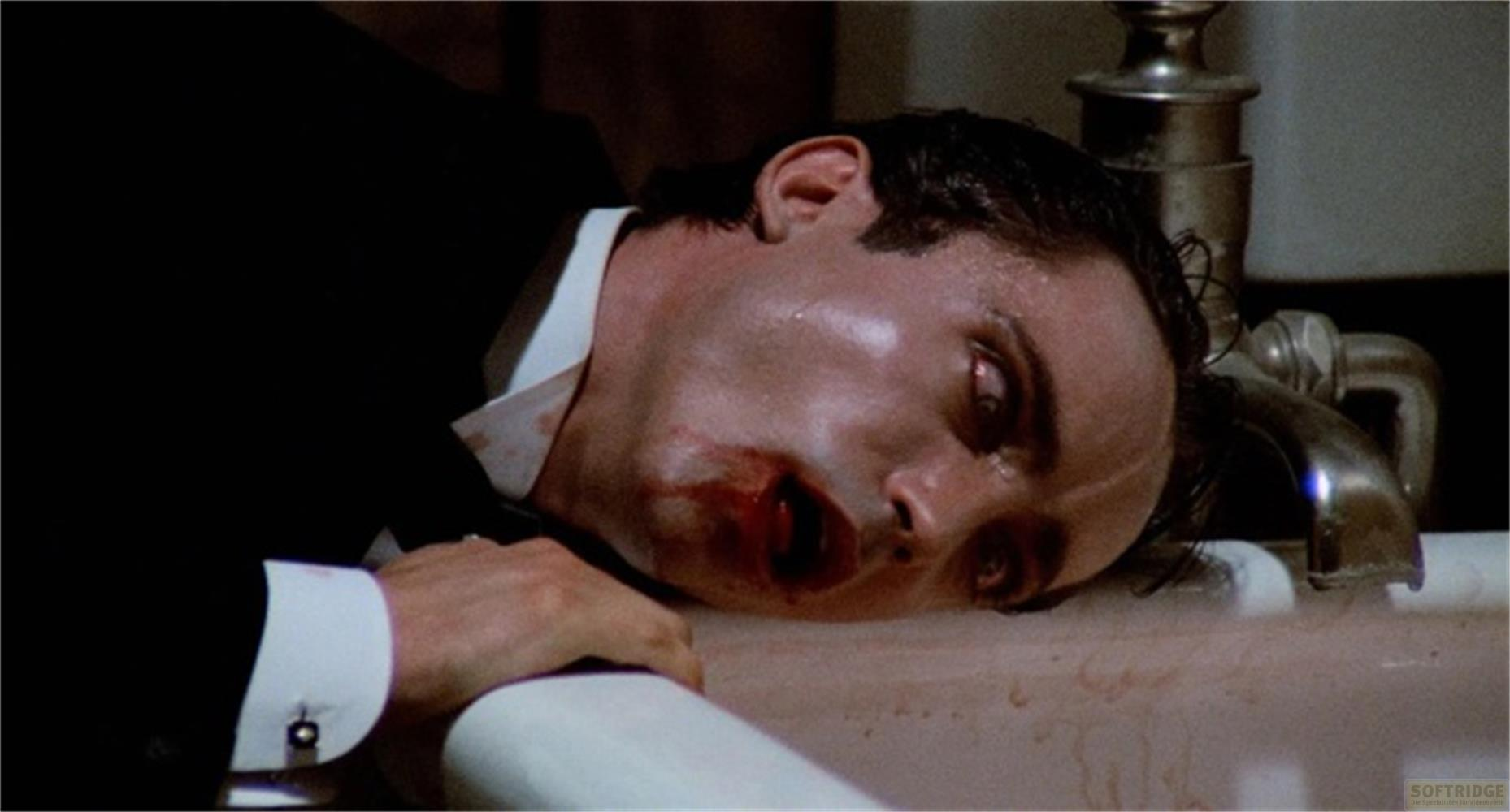 Uomo in abito sdraiato in una vasca da bagno, viso insanguinato, occhi aperti, che guarda verso l'alto.