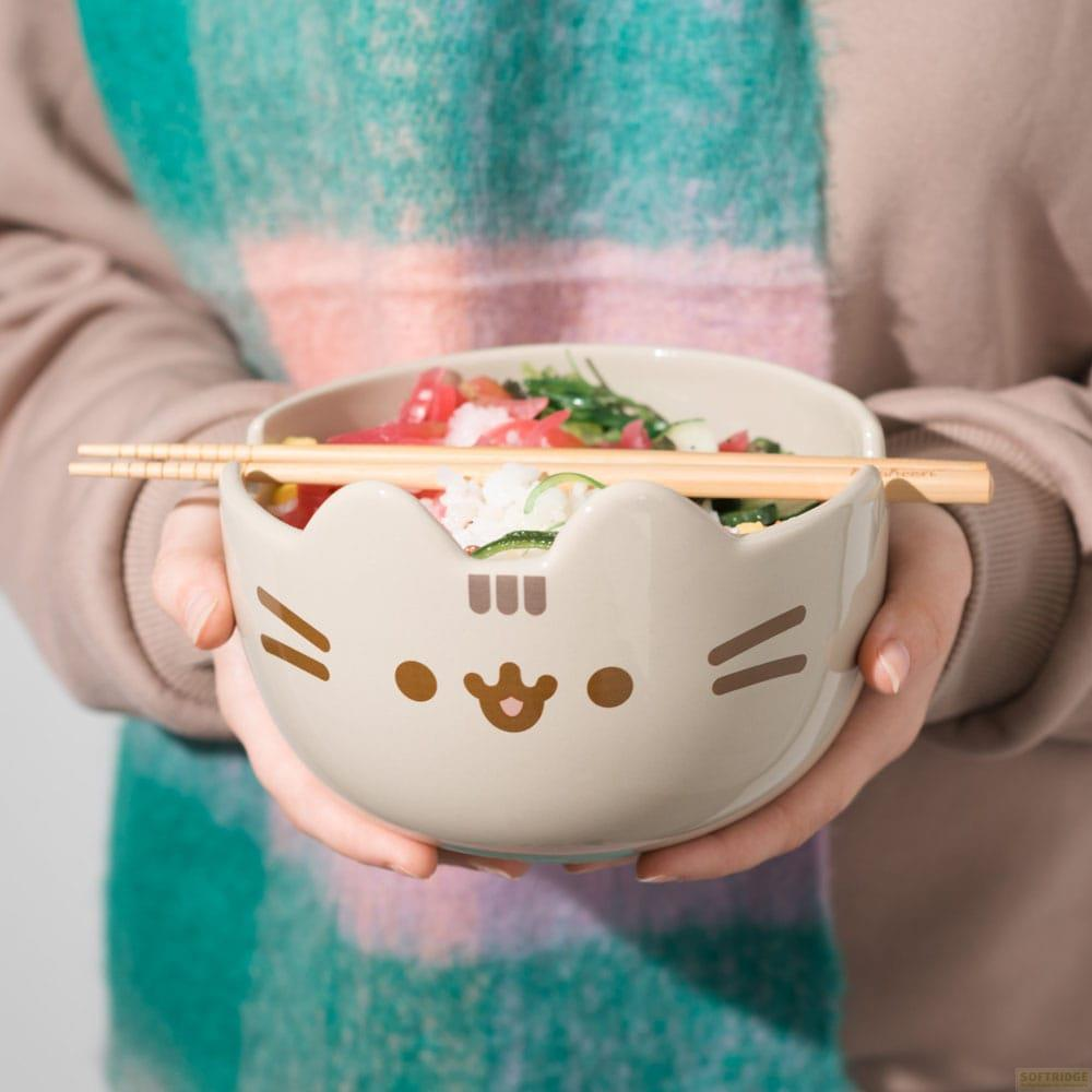 Una persona sostiene un cuenco con cara de gato lleno de arroz, pescado y verduras, con palillos encima.
