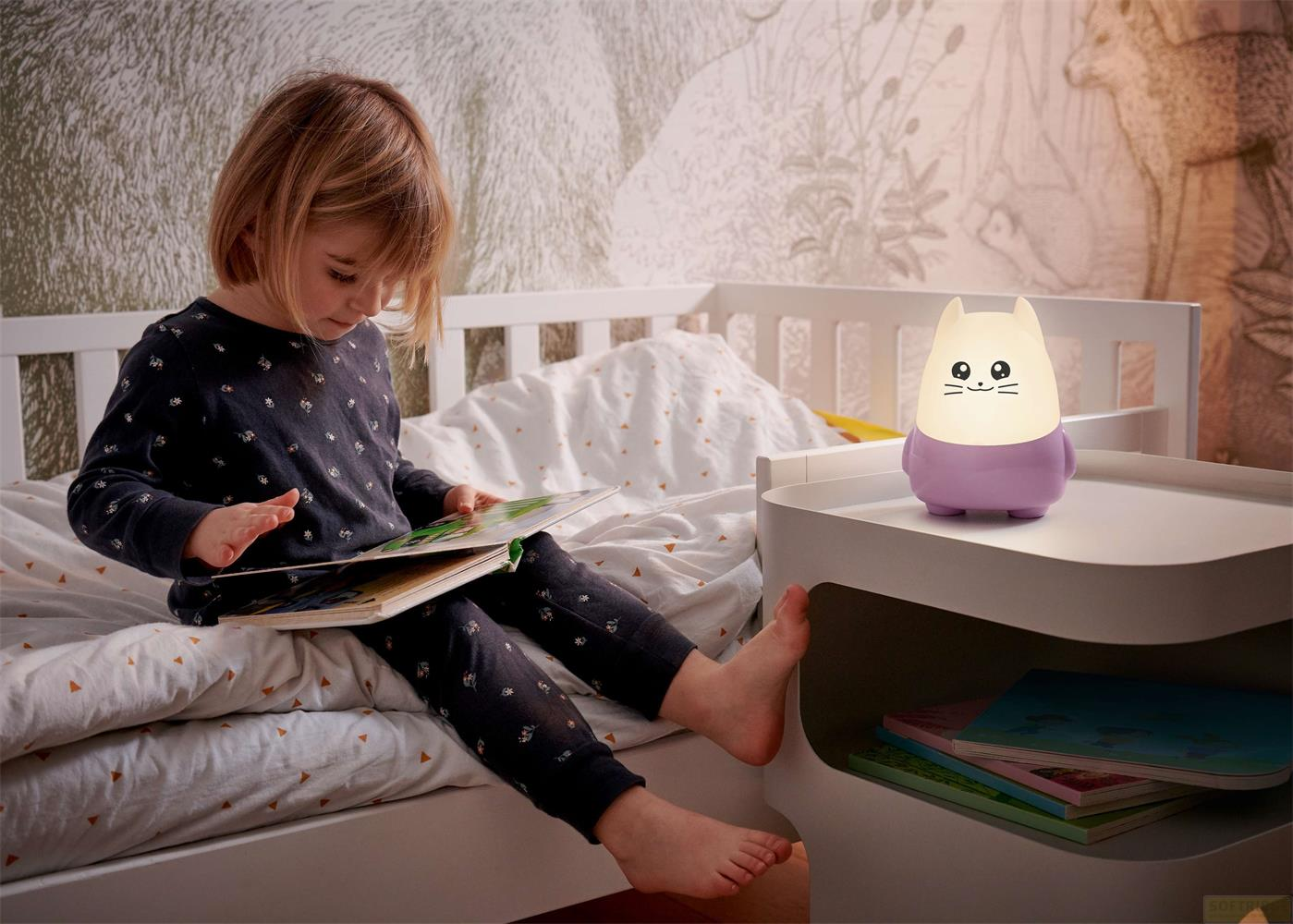 Une petite fille lit un livre au lit à côté d'une veilleuse chat.