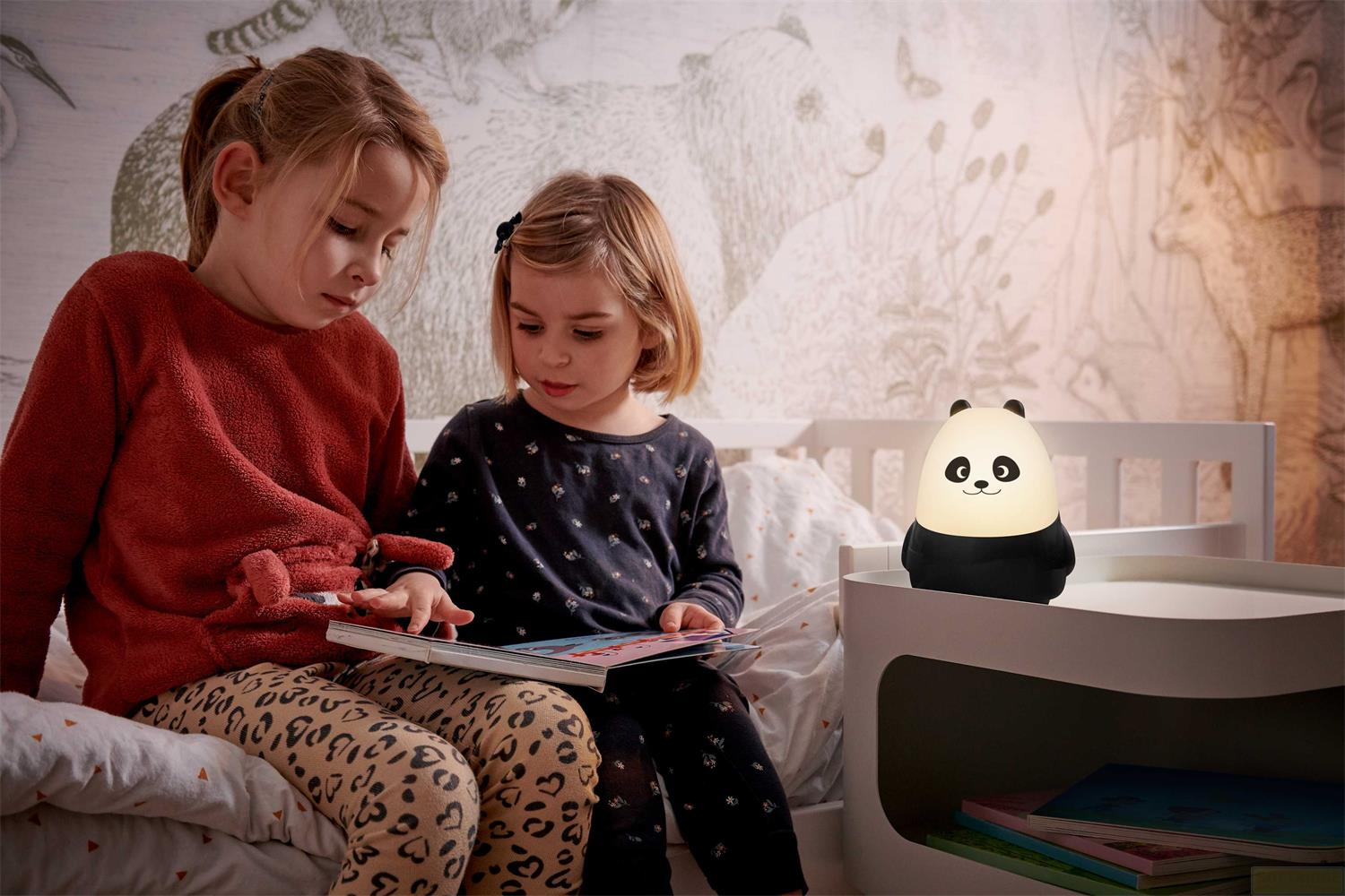 Deux filles assises sur un lit regardant un livre. Une veilleuse panda brille sur la table de chevet.