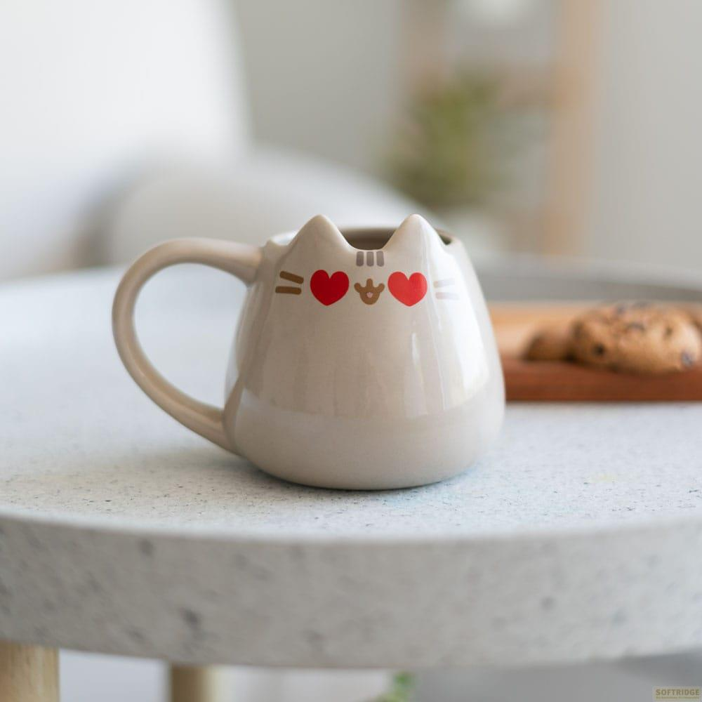 Una taza beige con orejas de gato y corazones rojos sobre una mesa.