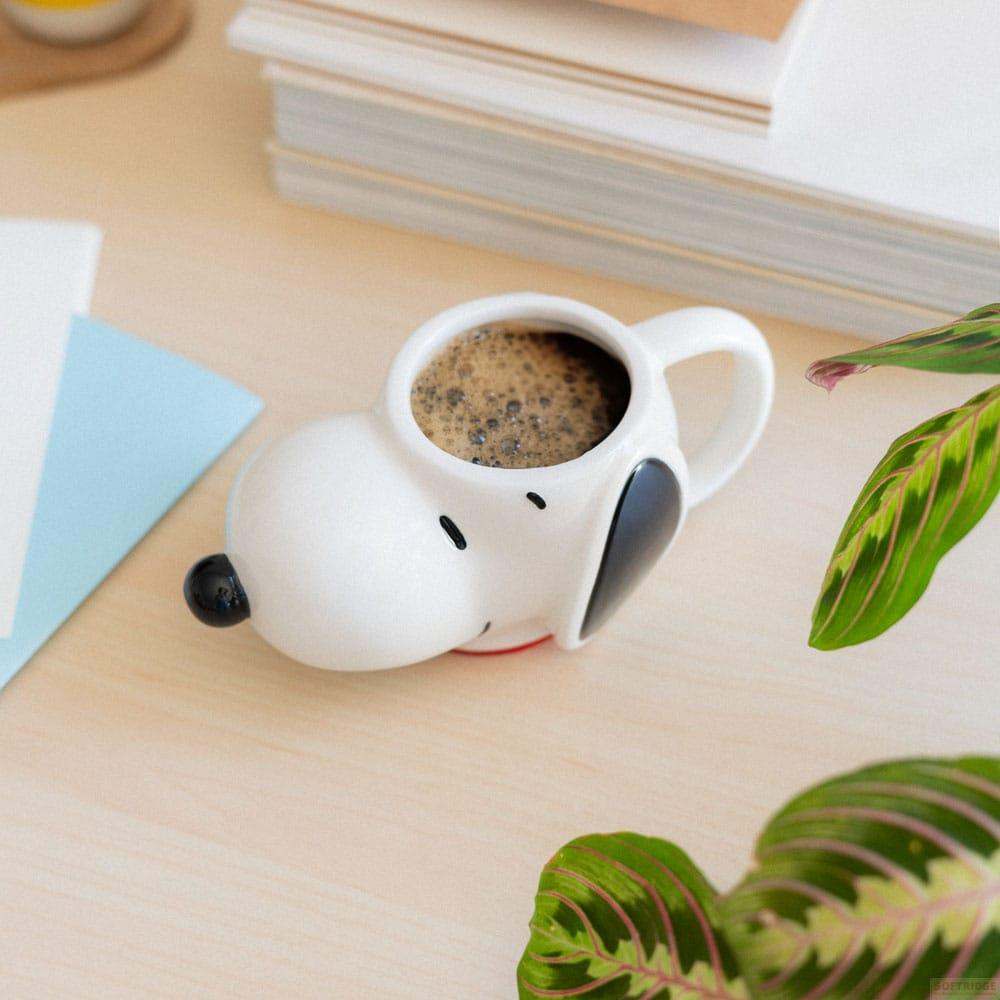 Una taza blanca de Snoopy con café se sienta sobre un escritorio de madera con libros y plantas.