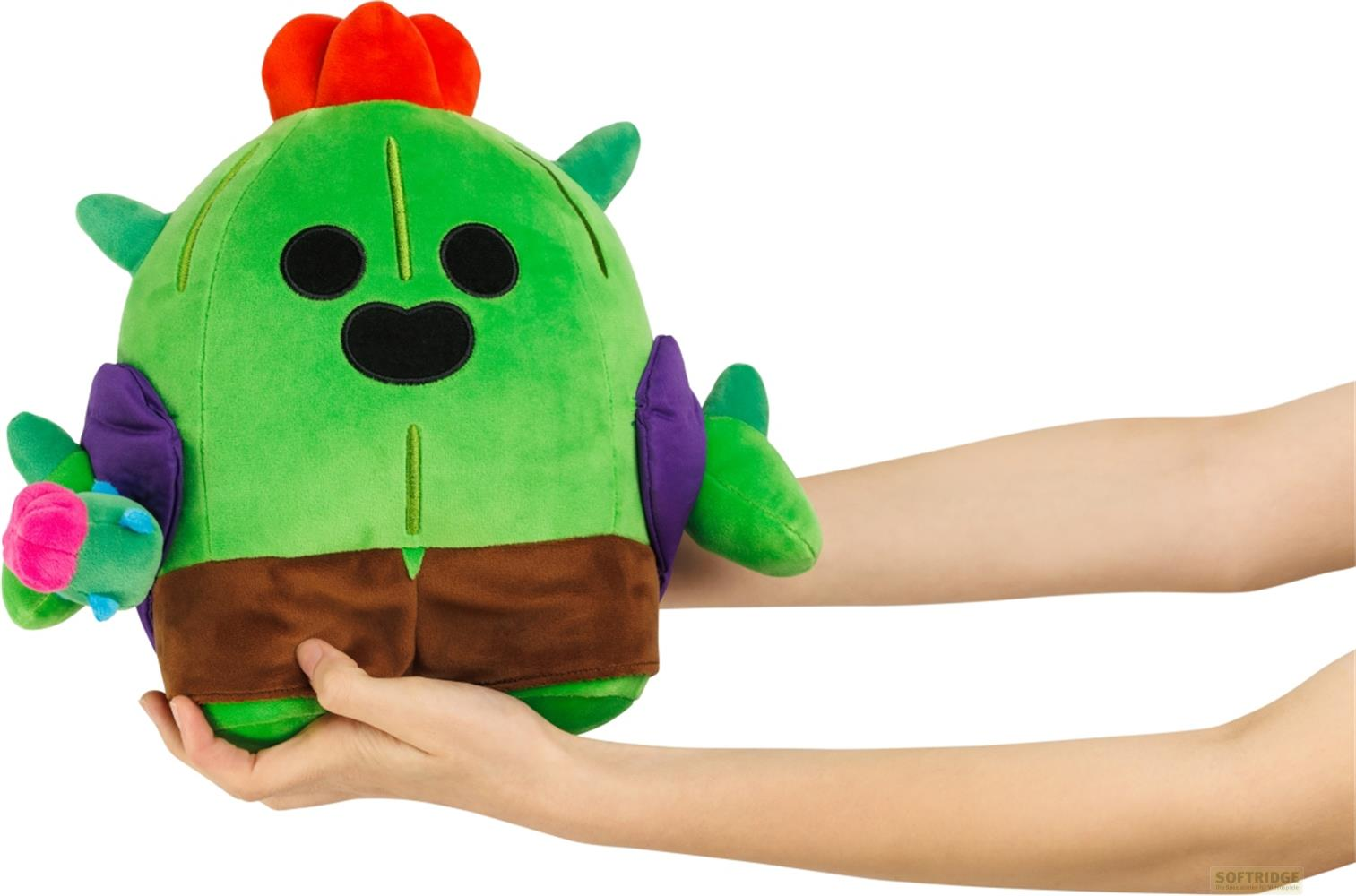 Peluche cactus verde con fiori rossi e pantaloncini marroni, tenuto da mani.