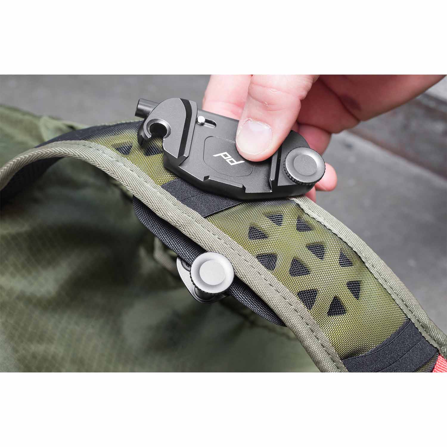 Una mano tiene una clip Peak Design grigia su una cinghia verde oliva.
