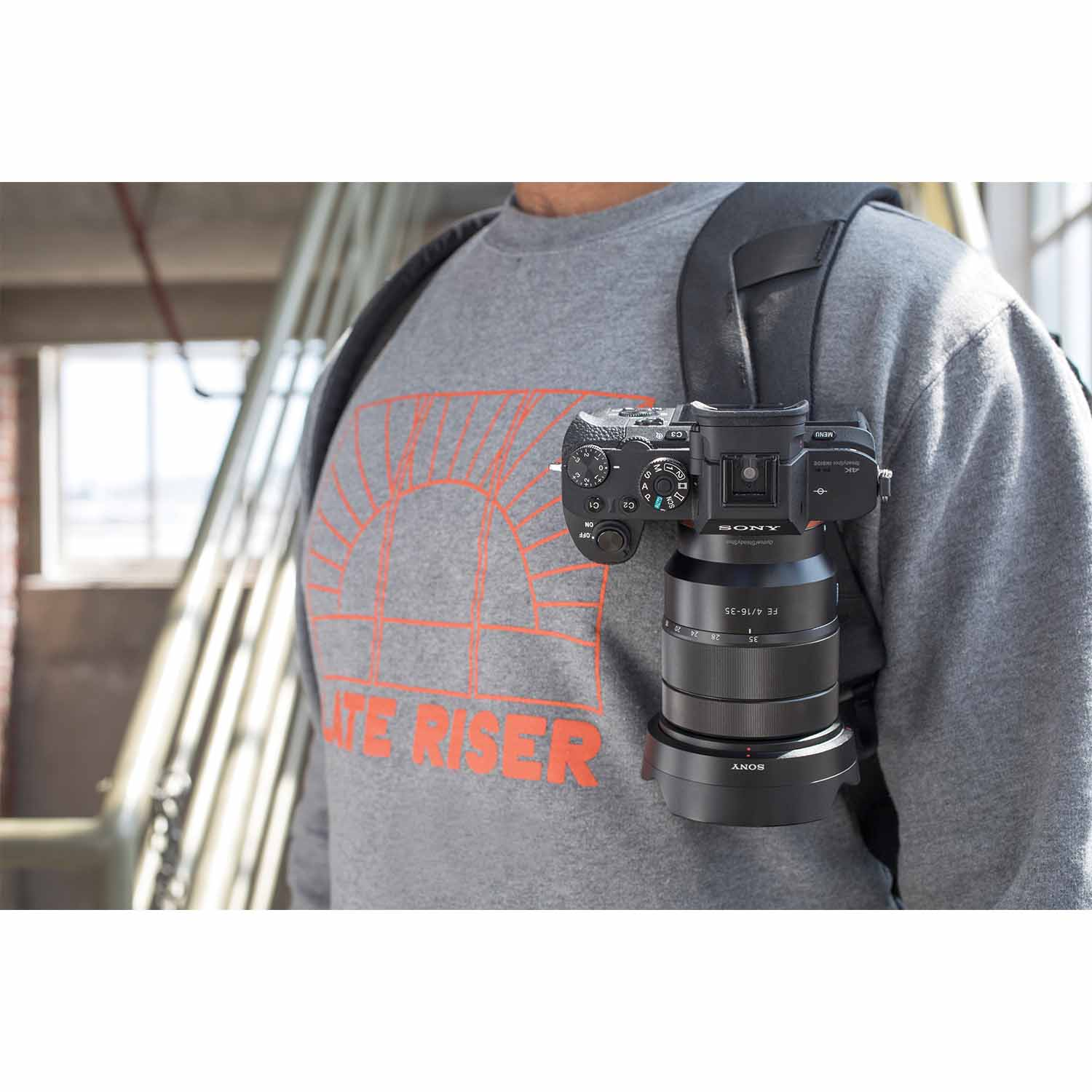 Un uomo indossa una fotocamera Sony con obiettivo sulla spalla. T-shirt grigia con stampa.