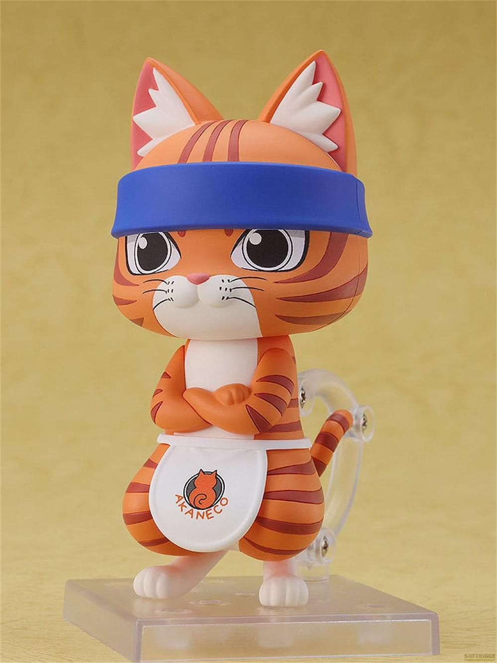 Eine orangefarbene Figur einer Katze mit blauer Stirnband und Schürze, steht auf Sockel.