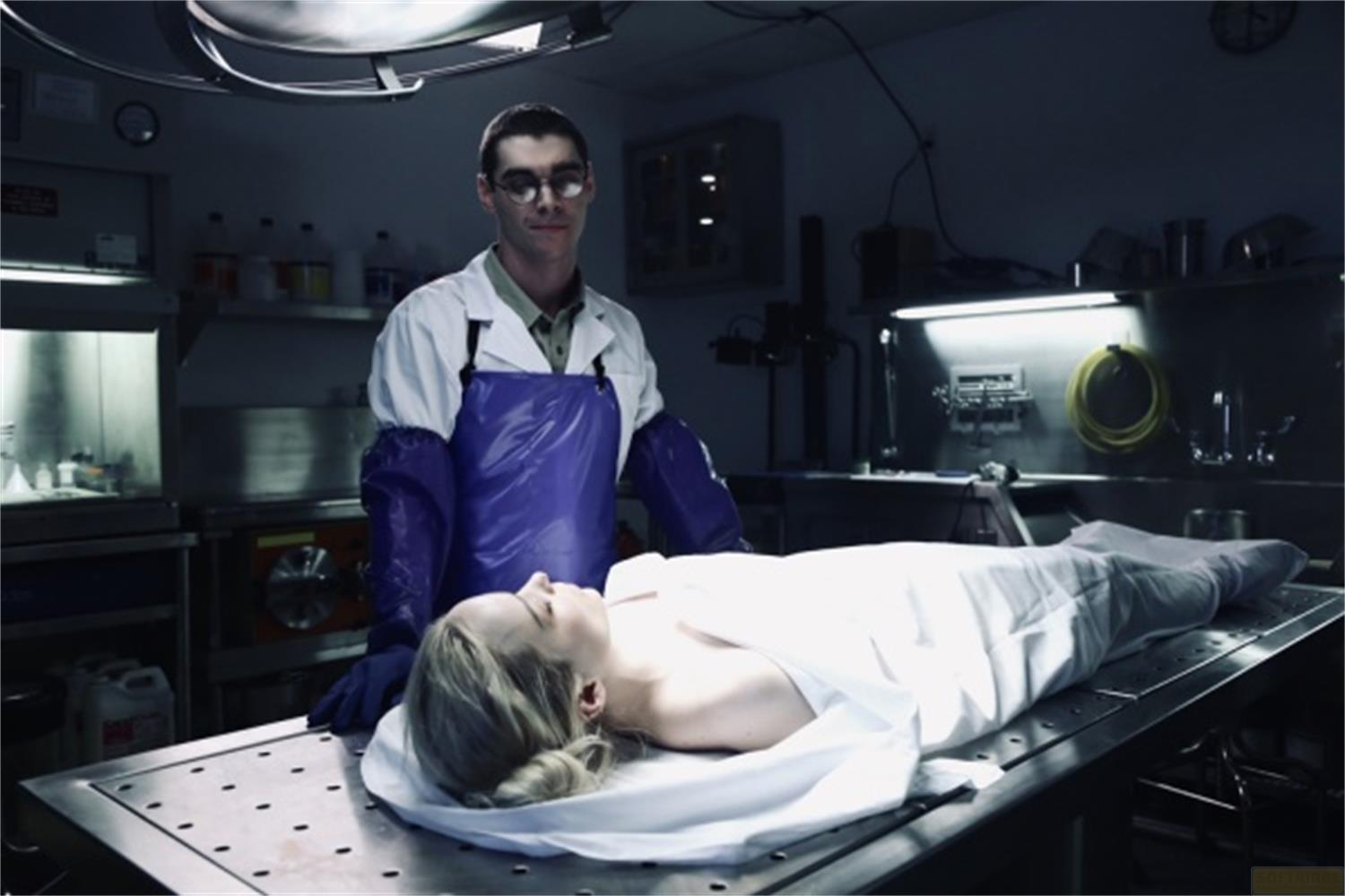 Un uomo in camice da laboratorio e grembiule blu sta sopra una donna su un tavolo.