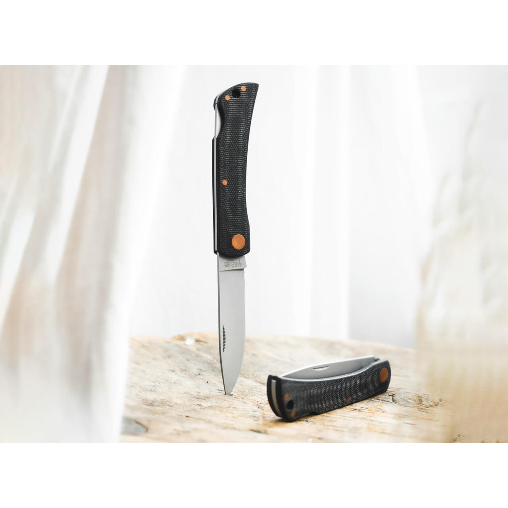 Ein Klappmesser mit schwarzem Griff und orangefarbenen Nieten liegt auf Holz.