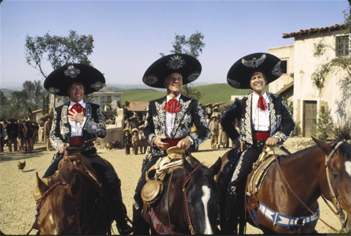 Tre uomini in abiti da mariachi cavalcano cavalli davanti a un villaggio messicano.