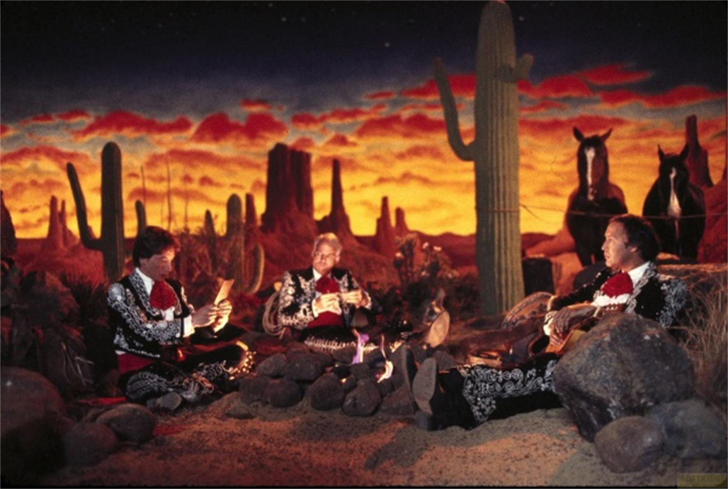 Tre uomini in abiti da mariachi seduti attorno a un falò nel deserto al tramonto.