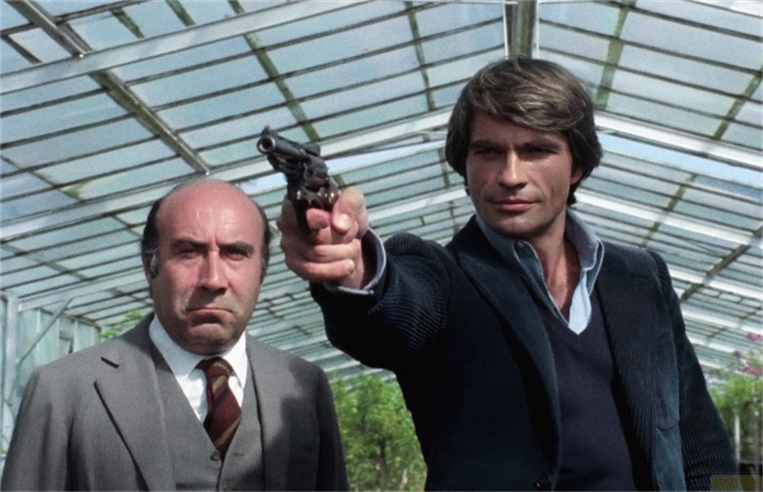 Un uomo in abito grigio e un uomo con pistola in una serra.