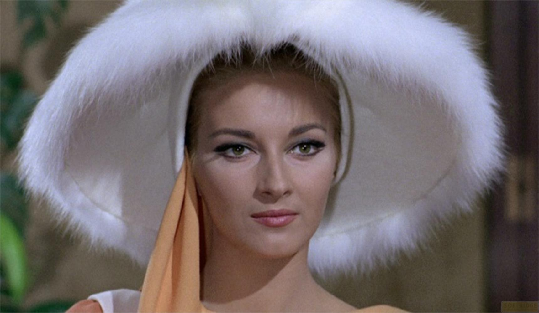 Una donna con trucco evidente indossa un grande cappello di pelliccia bianco e una sciarpa arancione.