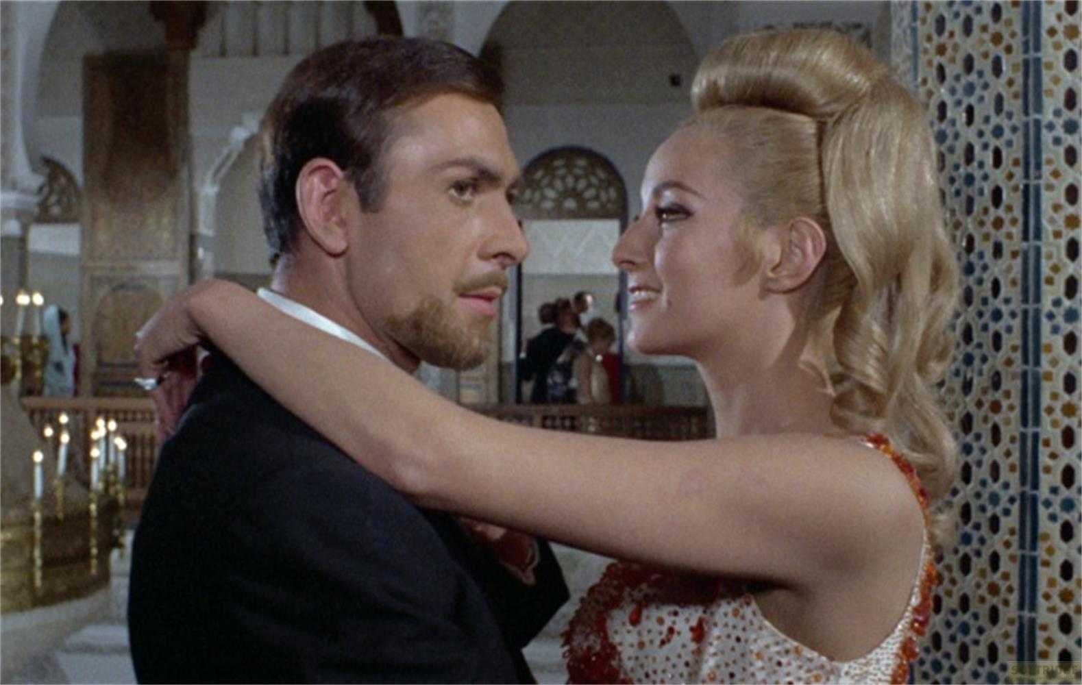 Un uomo con la barba e una donna bionda si abbracciano guardandosi.
