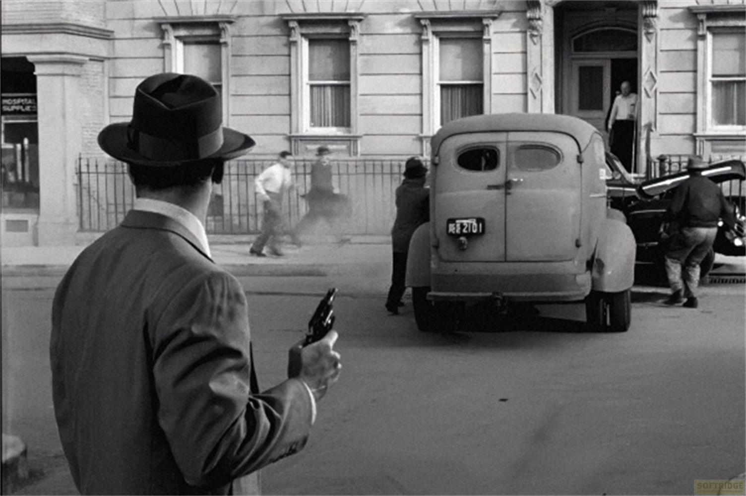 Un uomo con cappello tiene una pistola. Sullo sfondo, un furgone e altre persone.