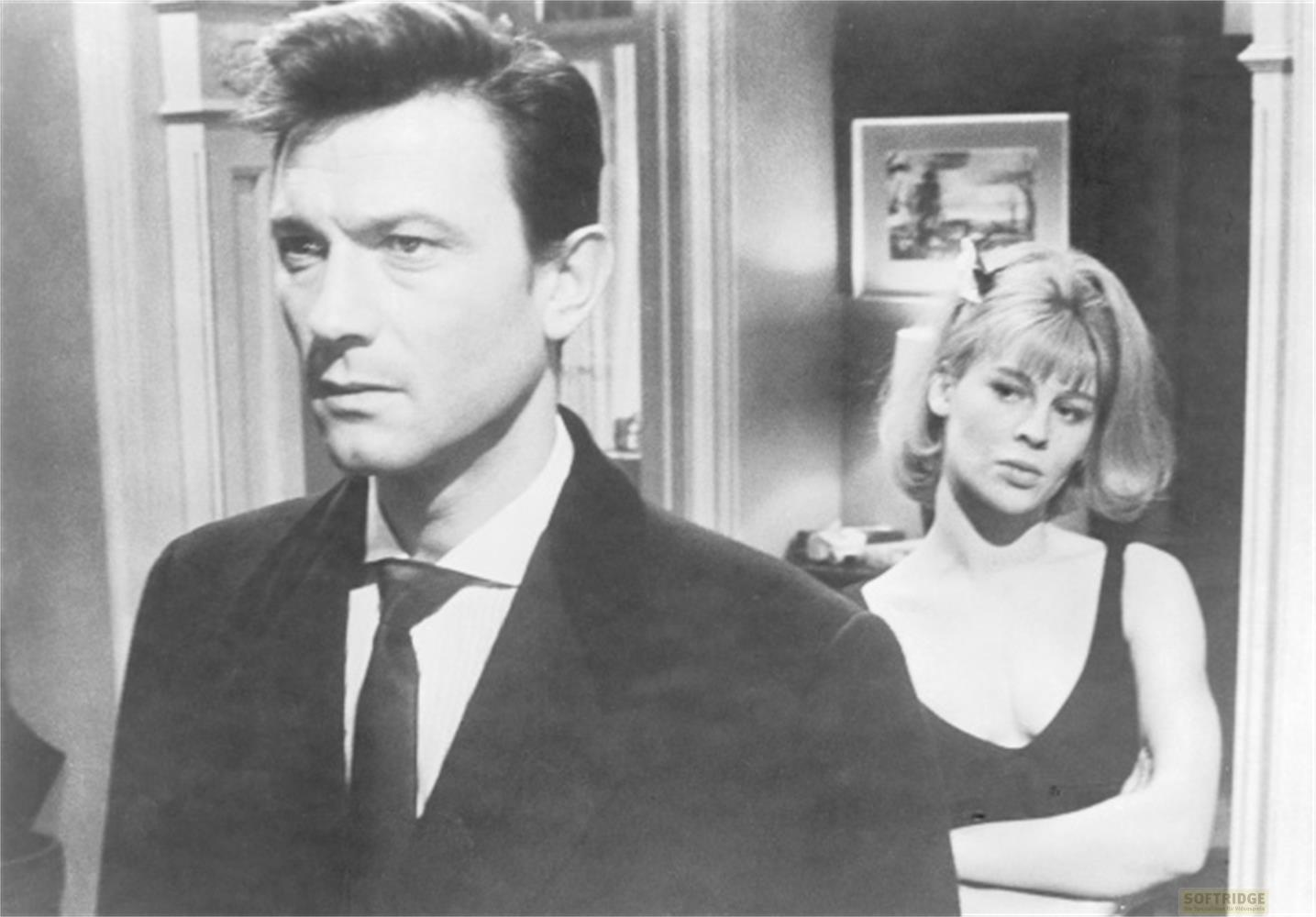 Uomo in abito e cravatta guarda a sinistra, donna in abito scuro dietro di lui con braccia incrociate.