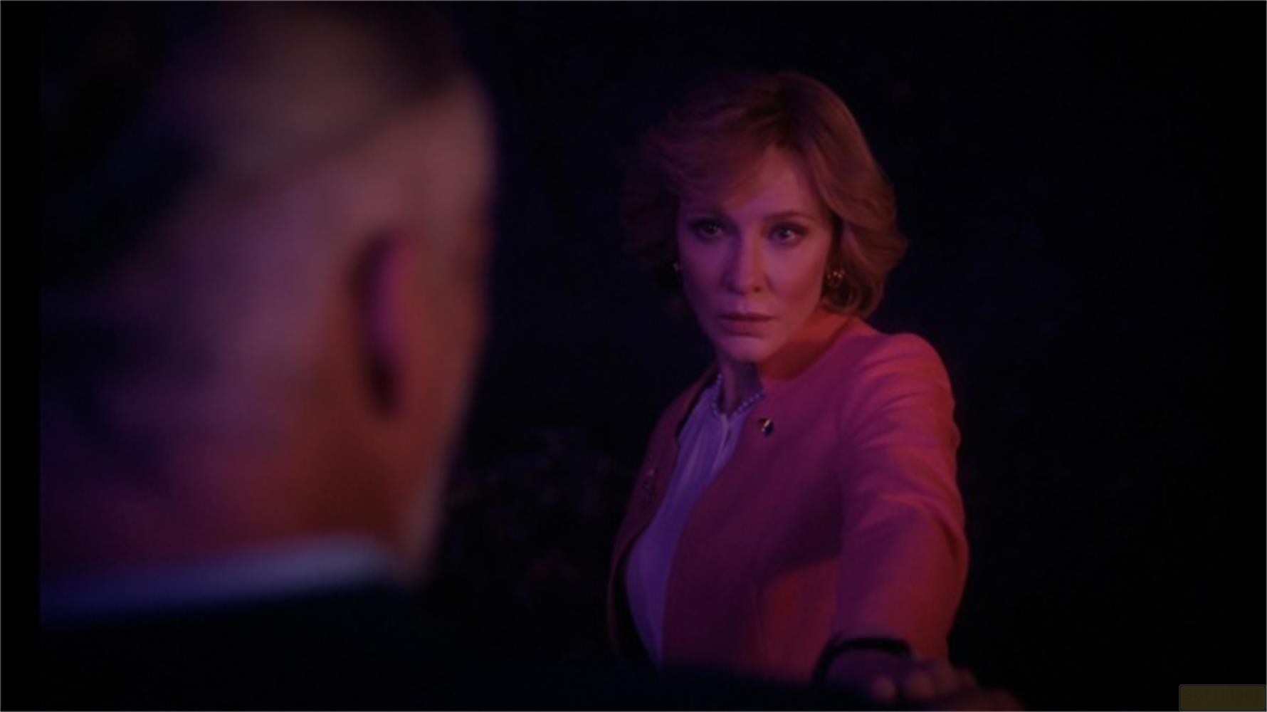 Una donna in blazer rosa guarda seriamente la telecamera, primo piano sfocato.