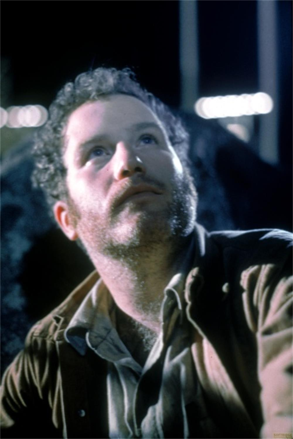 Gros plan d'un homme aux cheveux bouclés et à la barbe, regardant vers le haut. Il porte une veste marron sur une chemise claire.