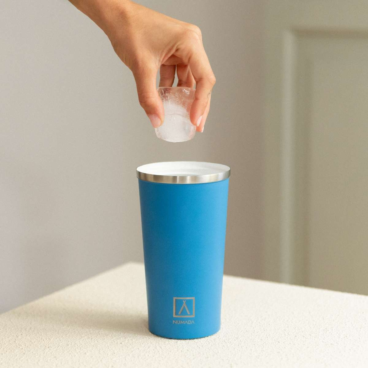 Una mano deja caer cubitos de hielo en un vaso azul con borde plateado.