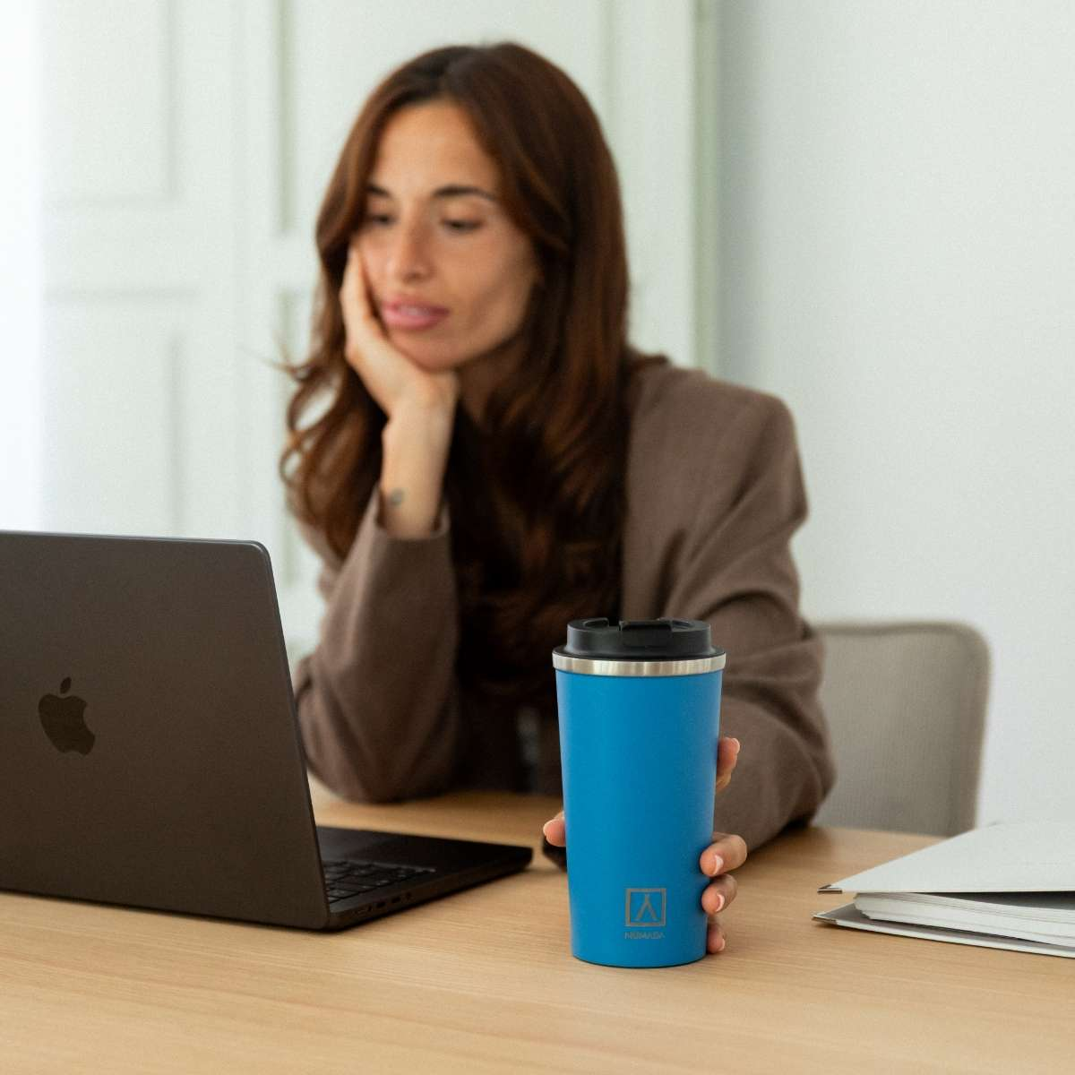 Una mujer en un escritorio sostiene una taza azul junto a un portátil.