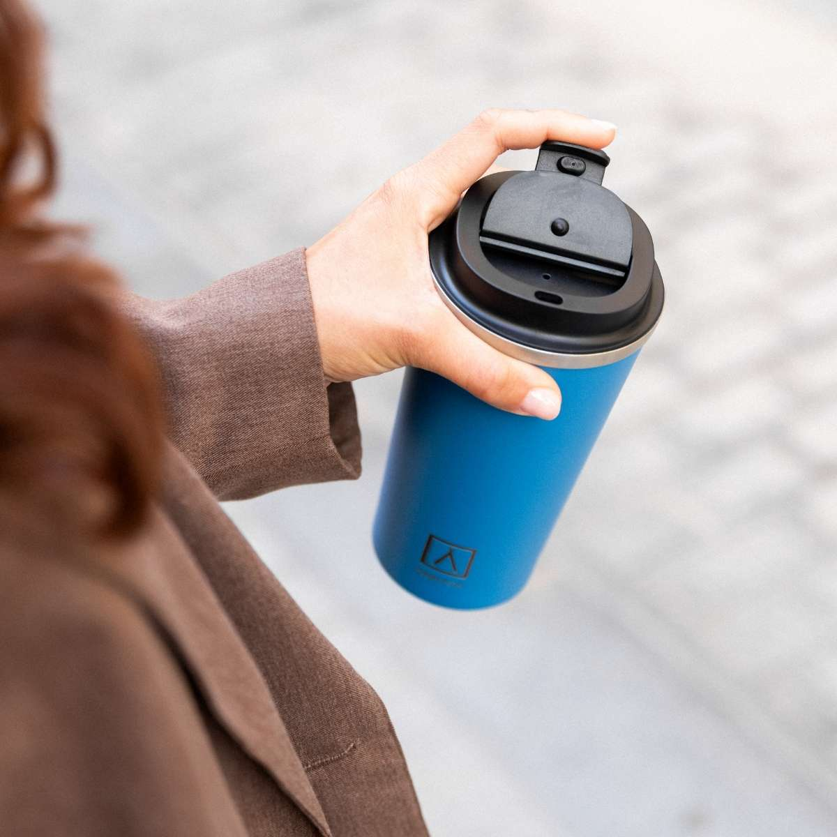 Una mano sostiene un vaso azul con tapa negra. Tiene un logo impreso.