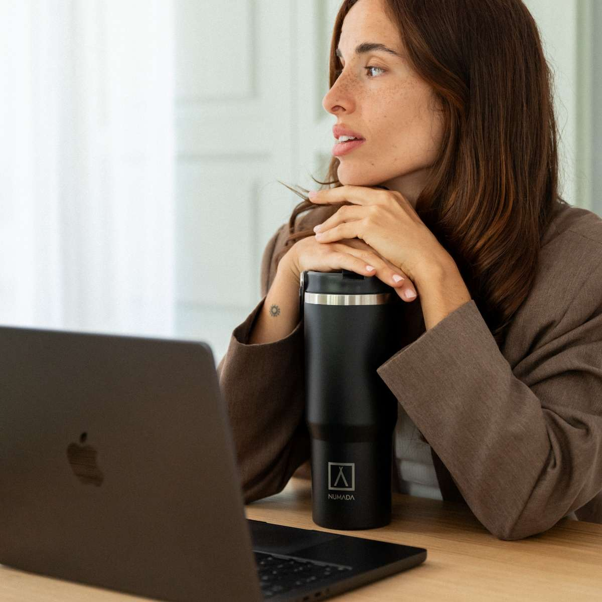 Una mujer pecosa mira lejos de una laptop, apoyando la barbilla en las manos cerca de un vaso negro.