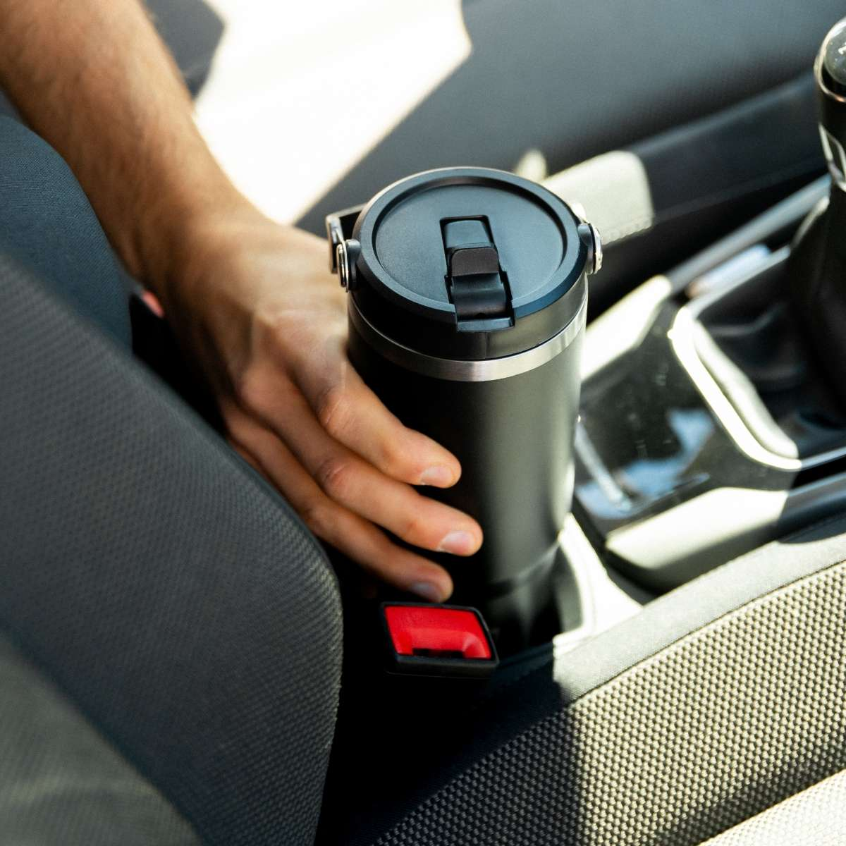 Una mano alcanza una taza de viaje negra en un portavasos de coche junto a un cinturón.