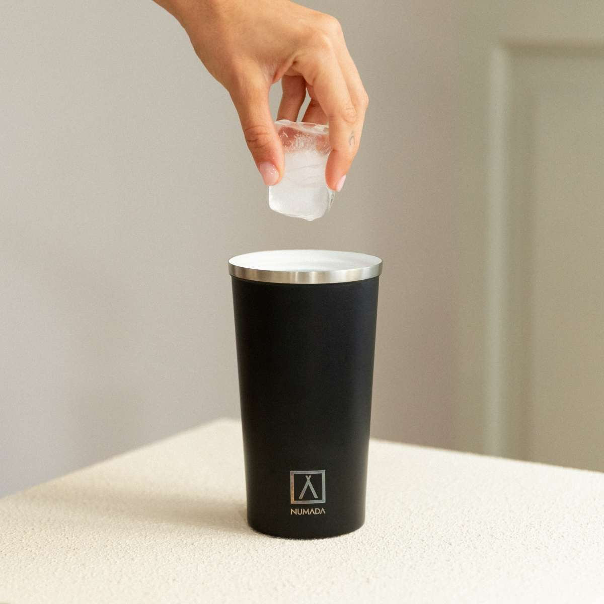 Una mano deja caer un cubito de hielo en un vaso negro.