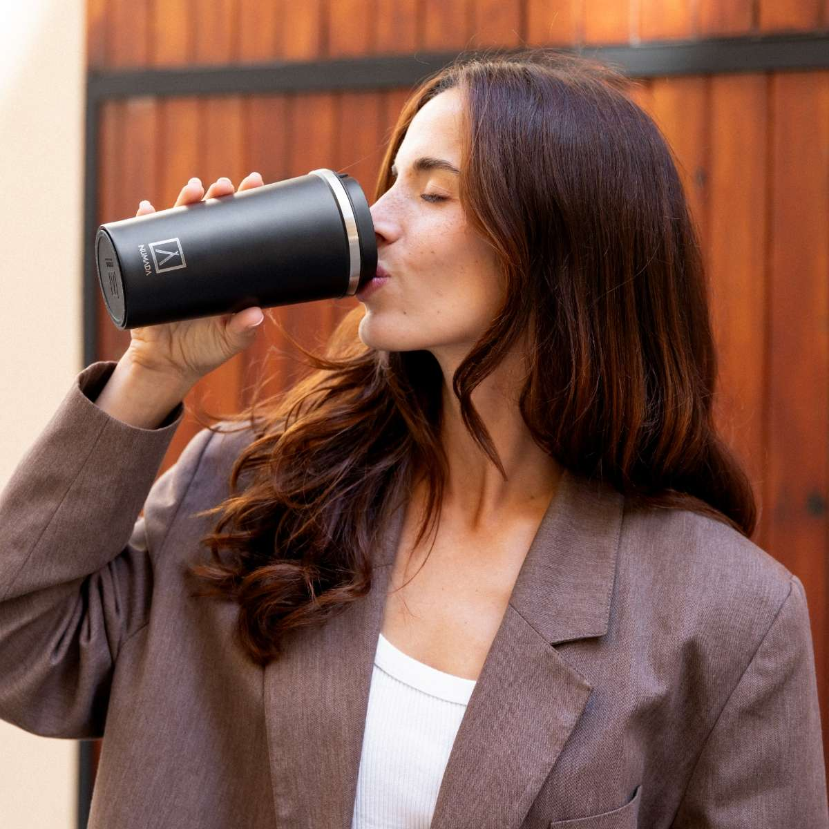 Una mujer de pelo castaño bebe de una taza negra frente a una puerta de madera.