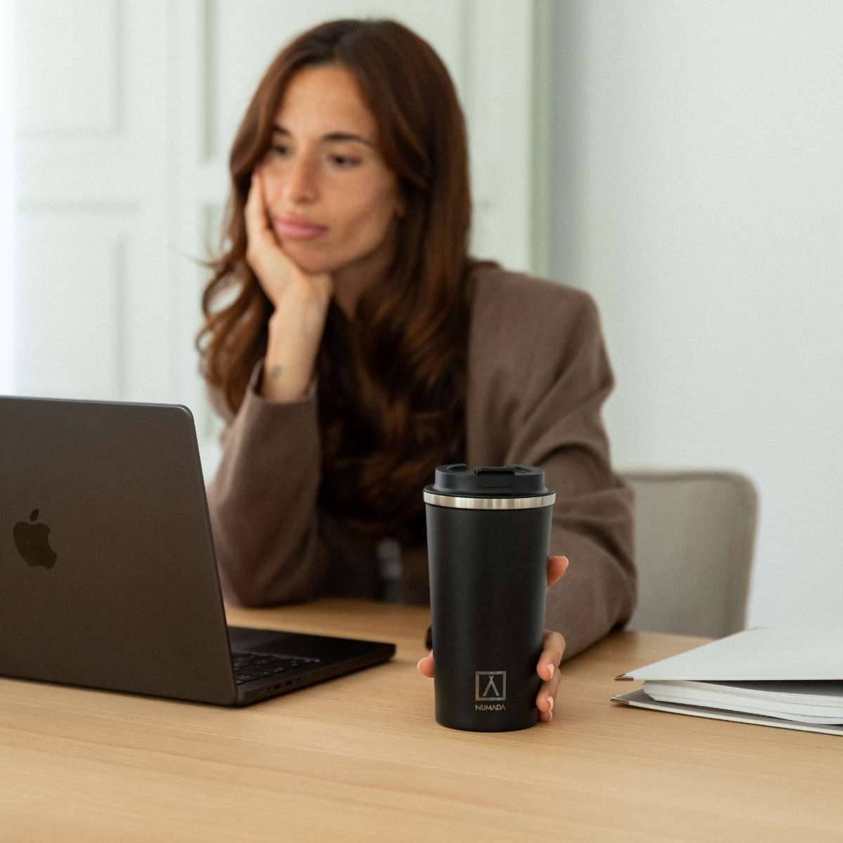 Una mujer de pelo castaño apoya la barbilla en la mano, mirando un portátil. Sostiene una taza negra en la otra mano.