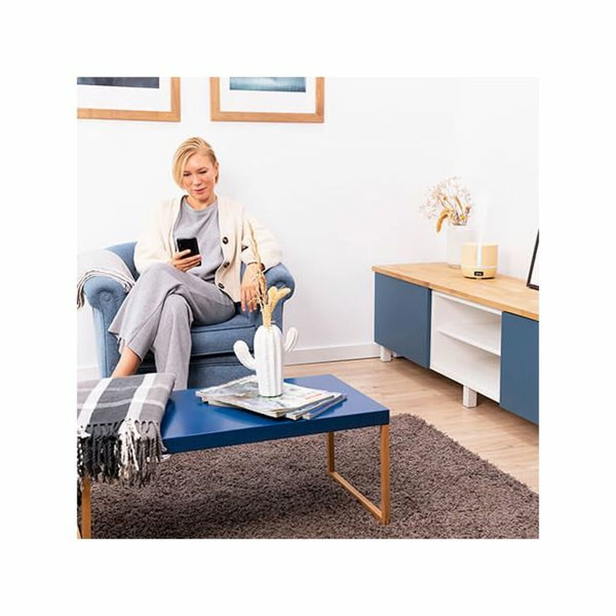 Kobieta siedzi w fotelu, patrząc na telefon. Przed nią stoi niebieski stolik kawowy.