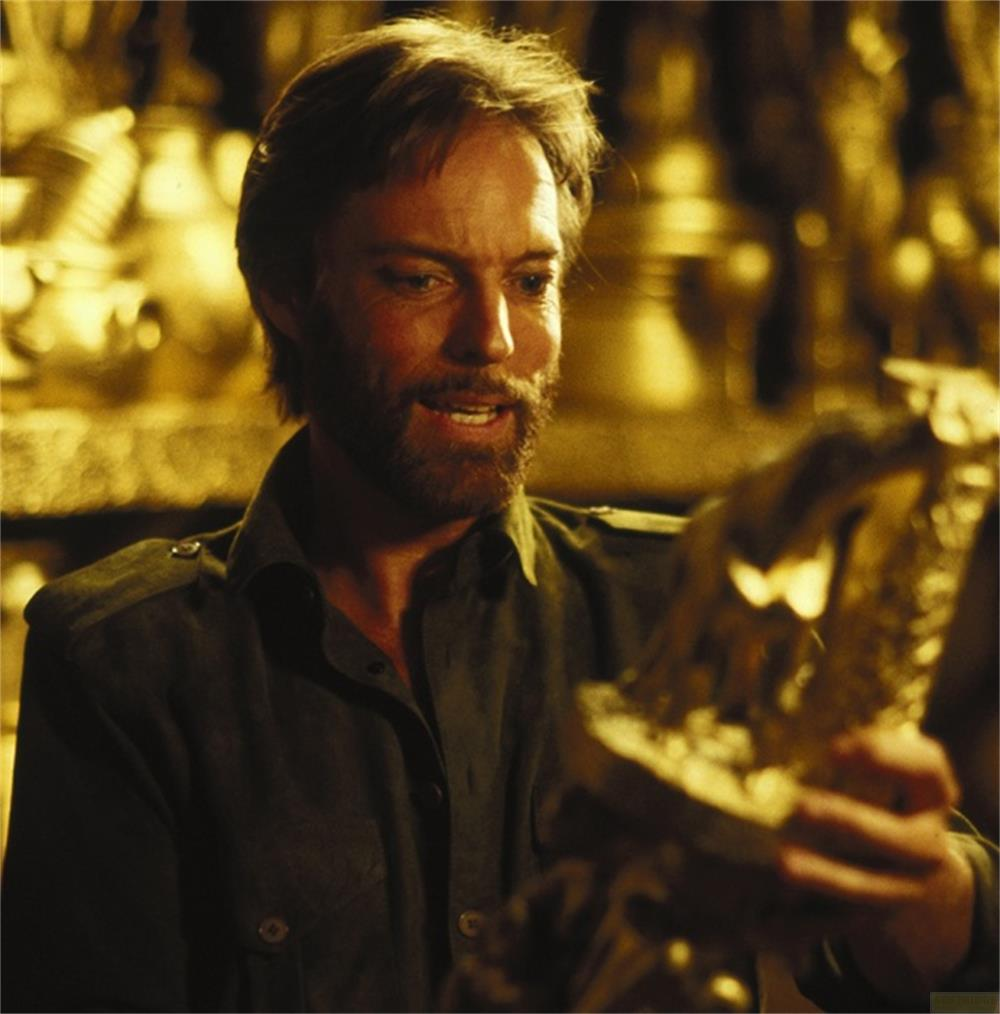 Homme barbu en chemise sombre tenant une statue d'aigle dorée sur fond doré.