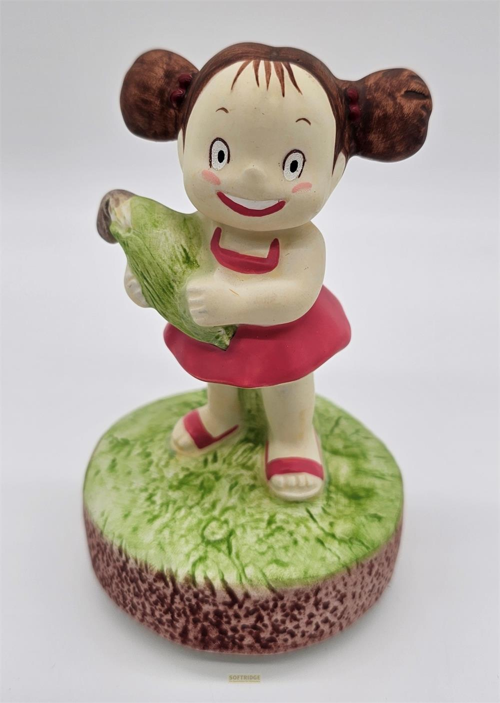 Figur eines Mädchens mit Duttfrisur, das ein grünes Bündel hält, auf einer Grasbasis.