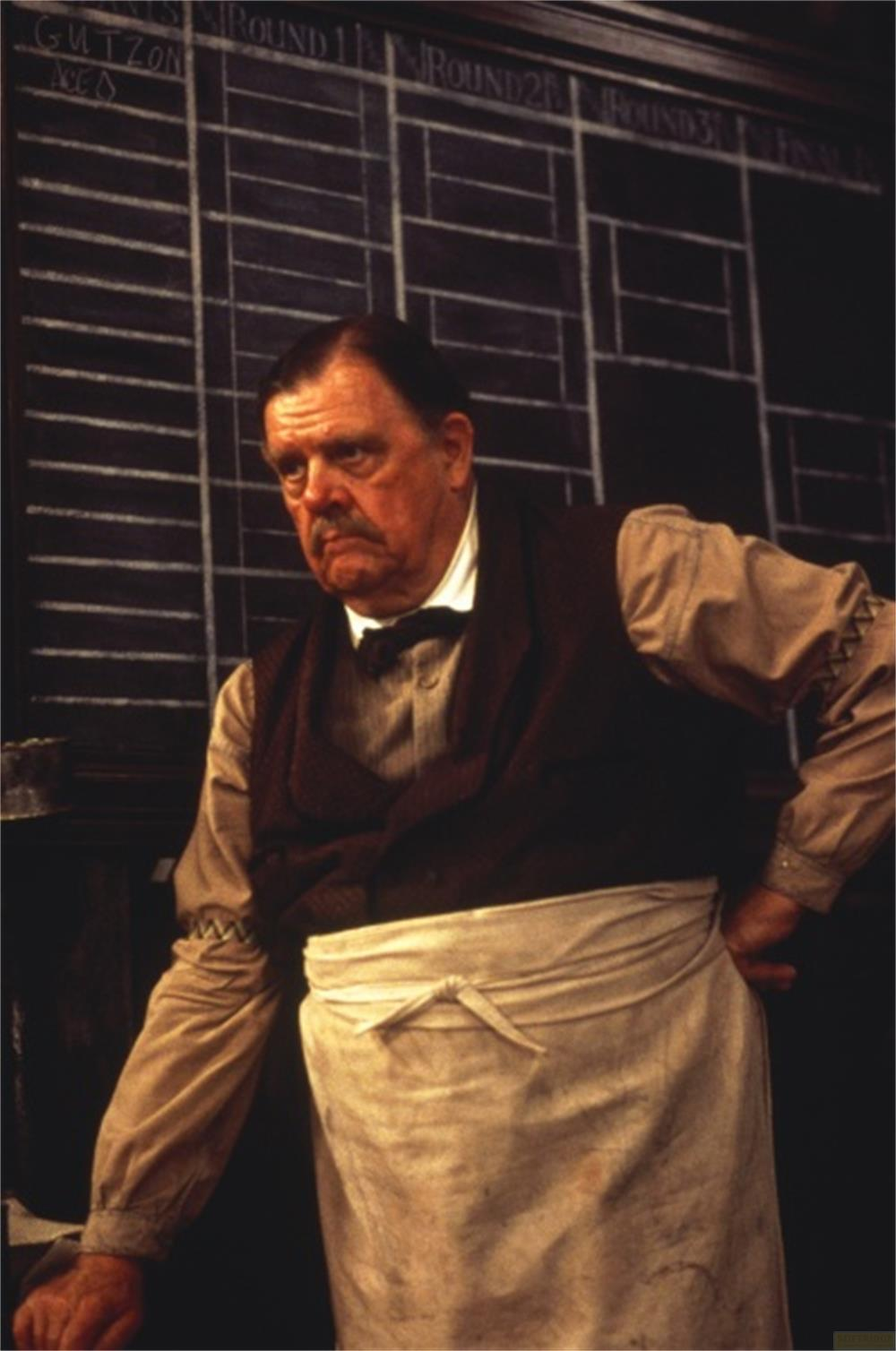 Un homme moustachu porte un gilet et un tablier blanc devant un tableau noir.