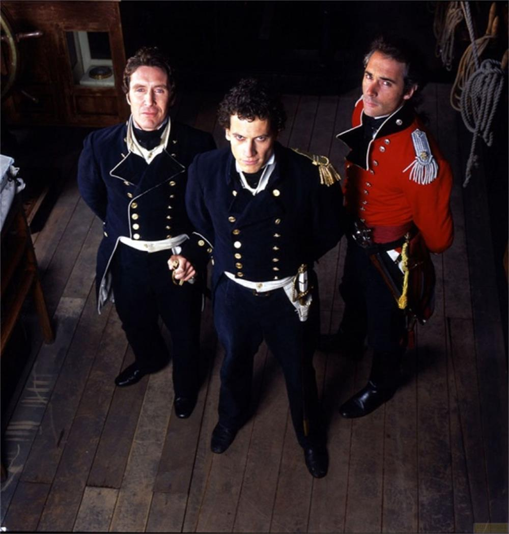 Tre uomini in uniformi storiche stanno su un ponte di legno. Due indossano blu scuro, uno rosso.