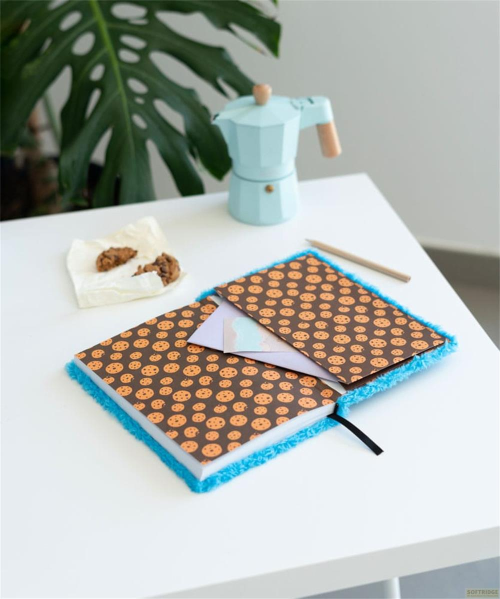 Un cuaderno abierto con estampado de galletas, al lado galletas, una cafetera y un lápiz.