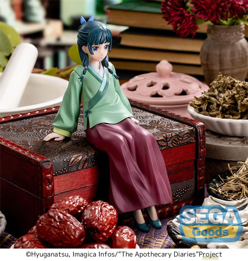 Une figurine assise sur un coffre orné, entourée d'herbes et de livres.