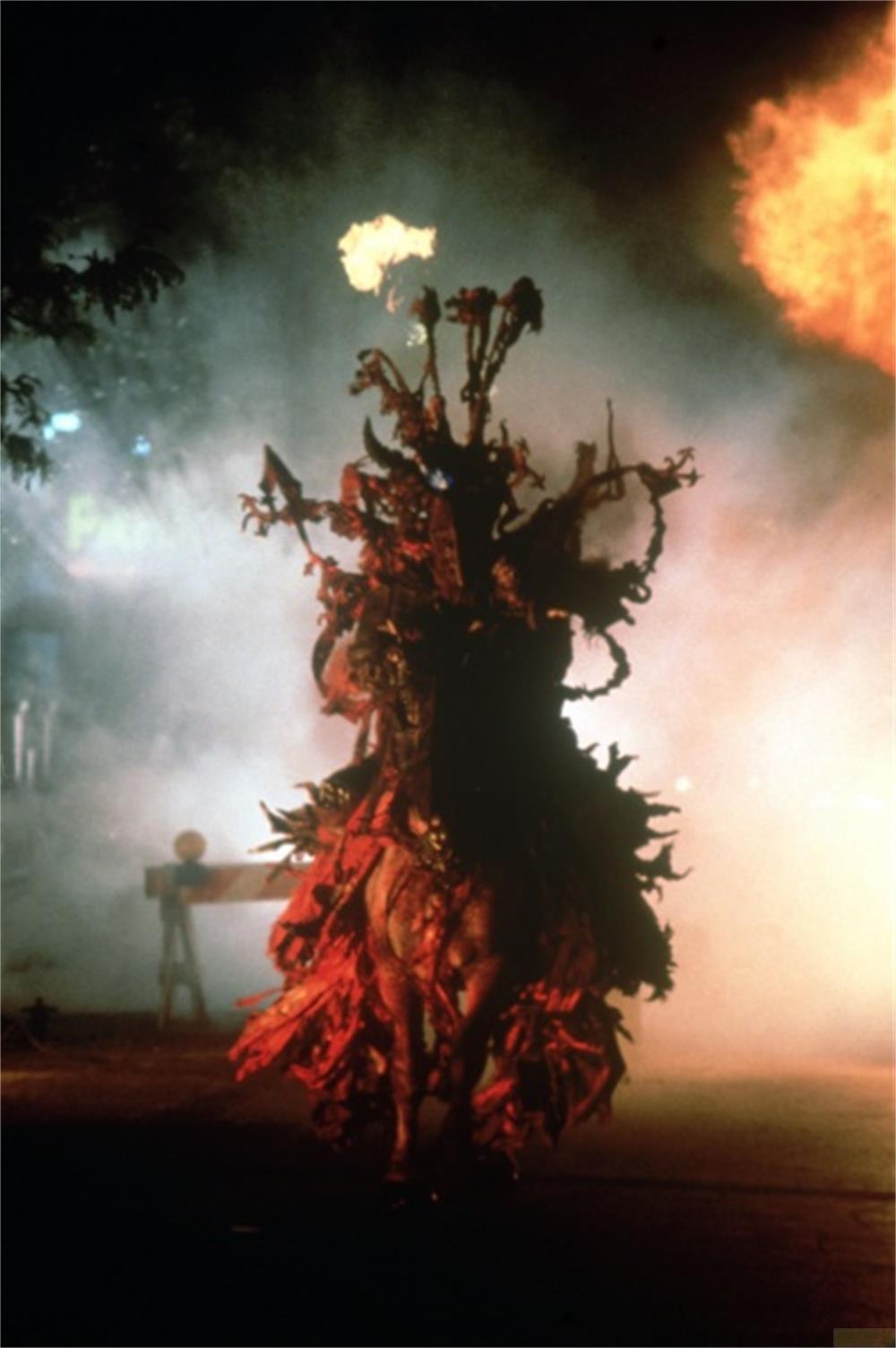 Une créature en costume rouge se dresse dans la fumée devant des flammes.
