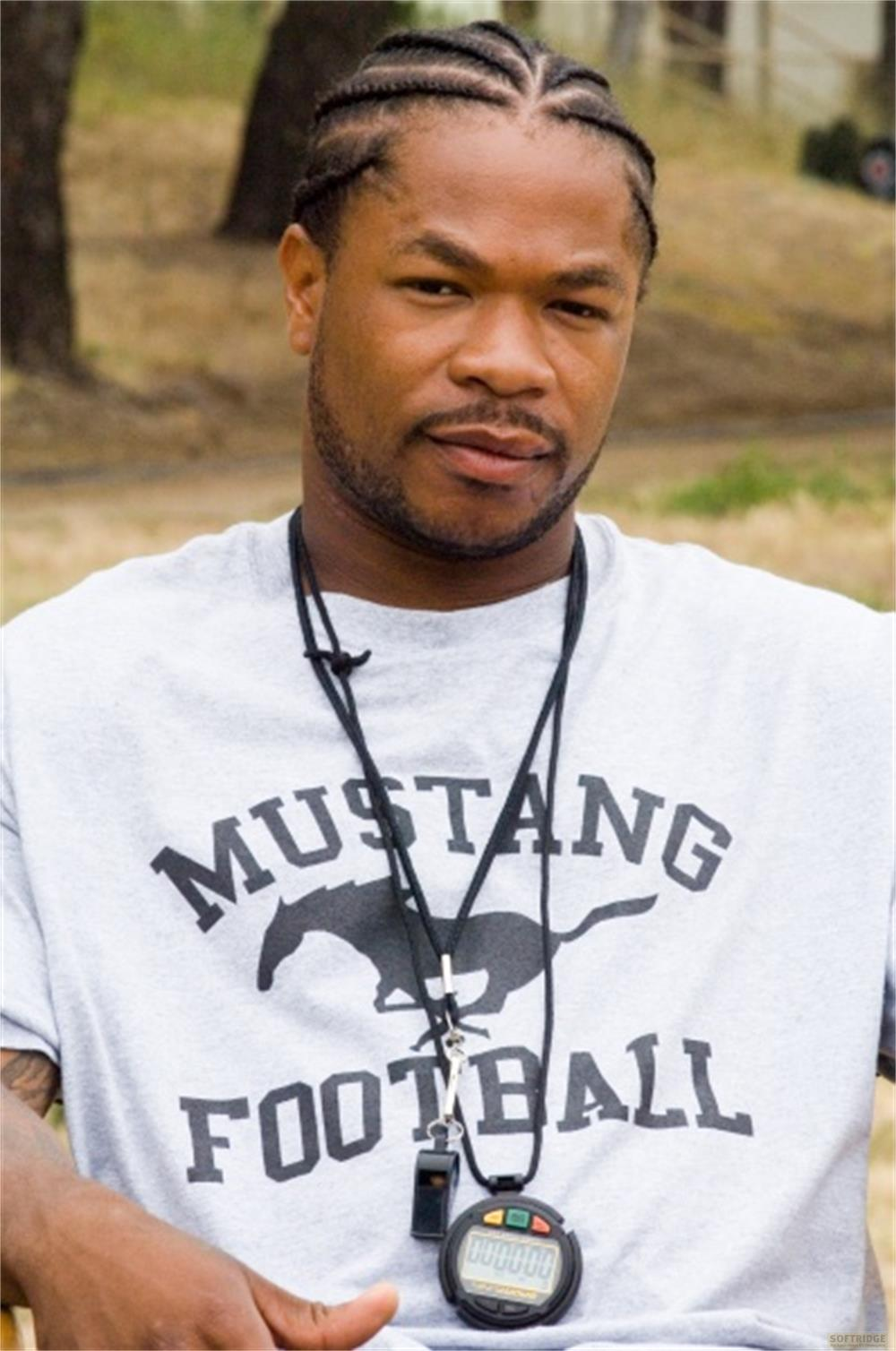 Ein Mann mit geflochtenen Haaren trägt ein "Mustang Football" T-Shirt und eine Stoppuhr.