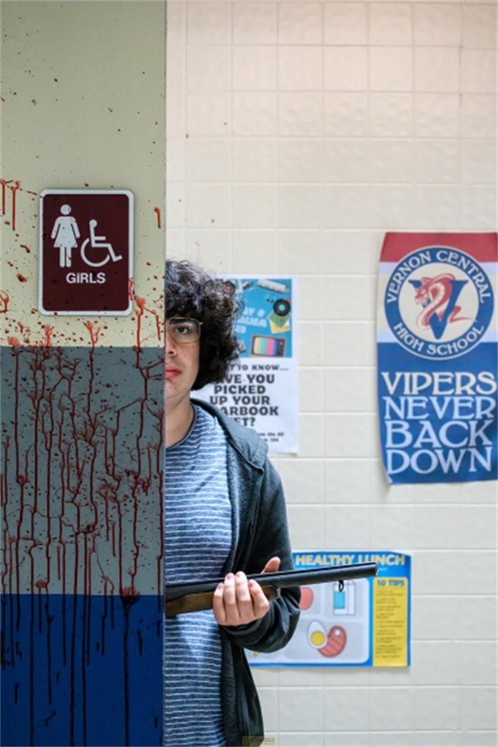 Un ragazzo con occhiali e fucile si trova davanti a un muro macchiato di sangue e poster scolastici.