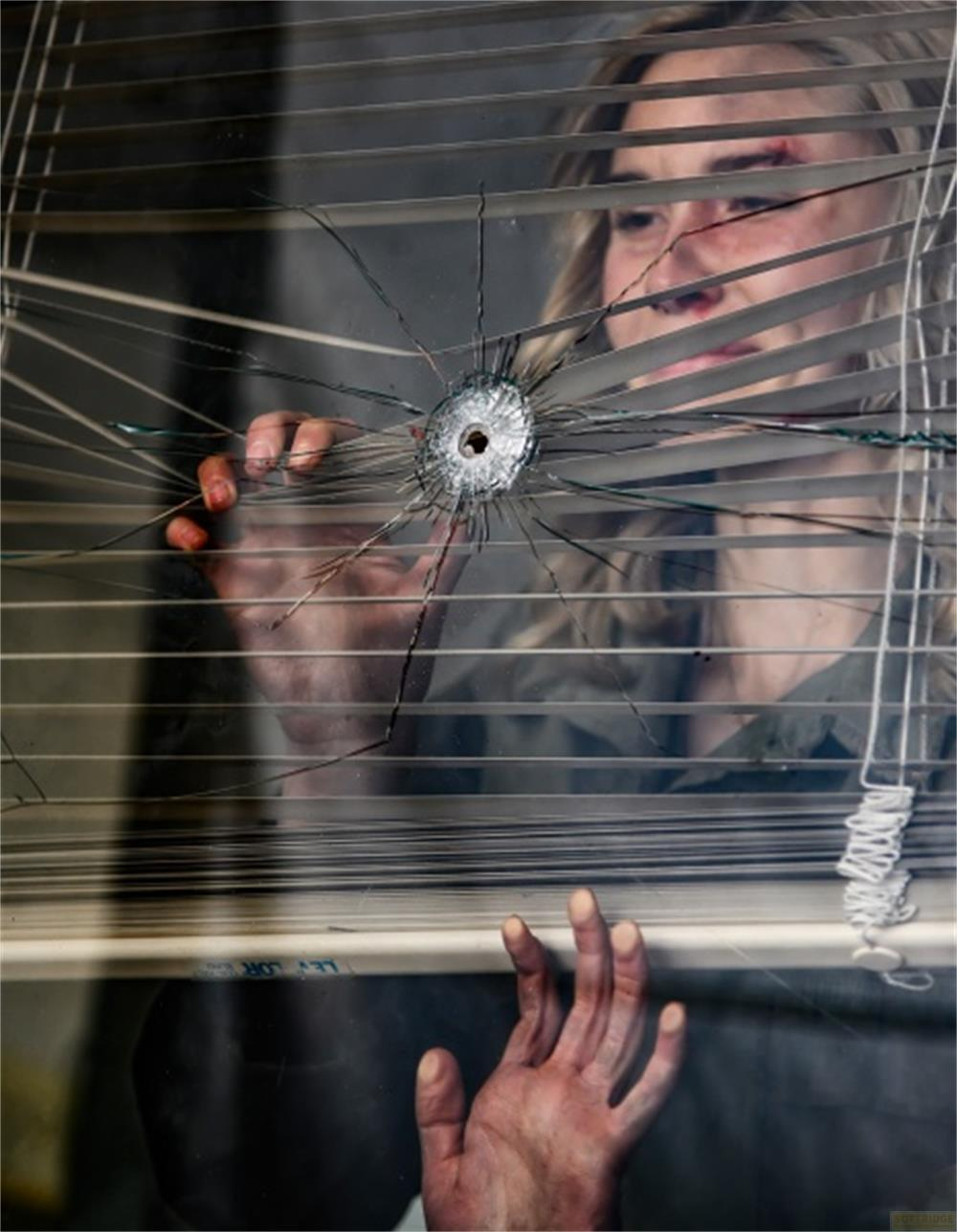 Una donna ferita guarda attraverso una finestra crivellata di proiettili con tende.