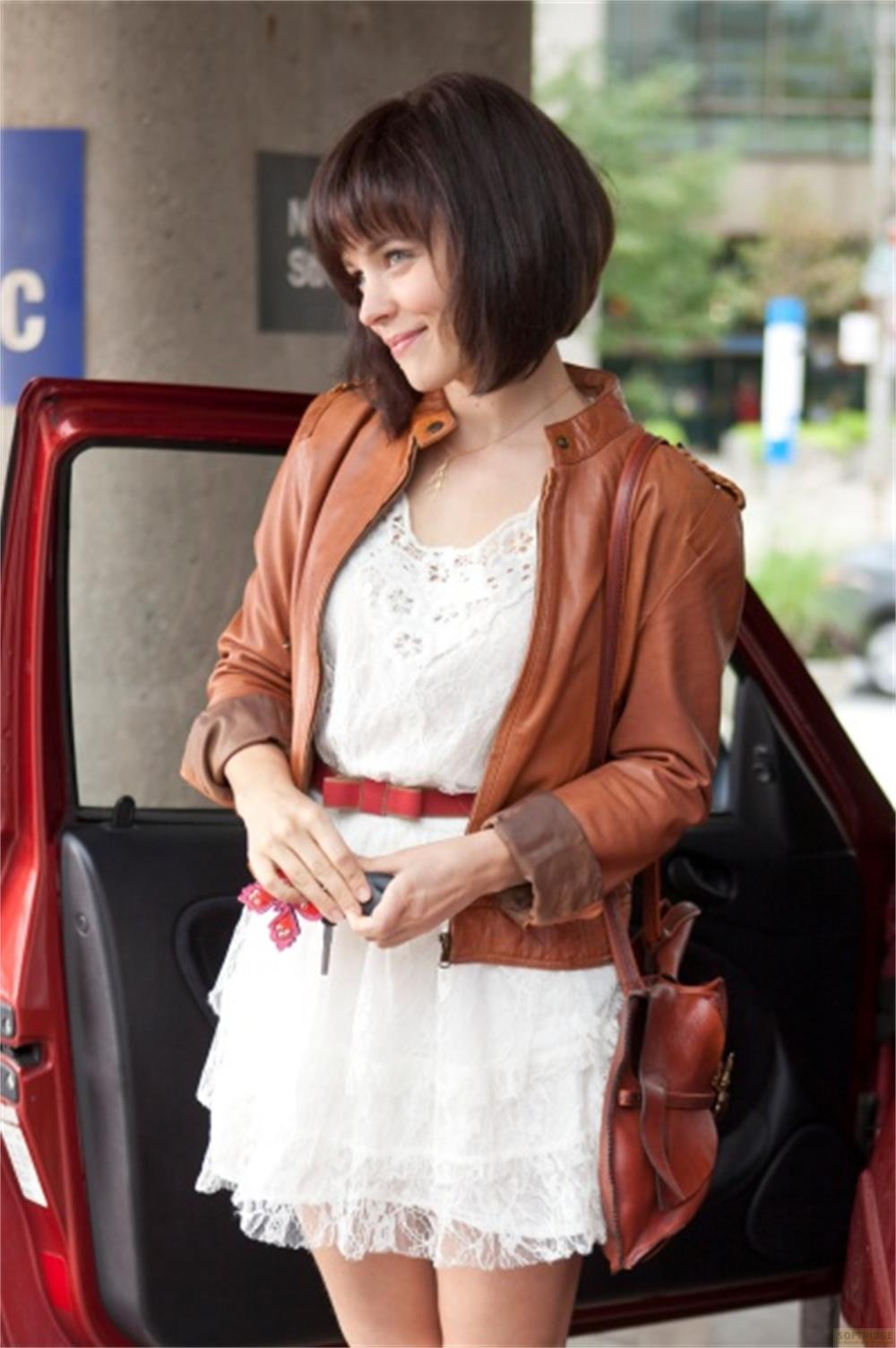 Femme en blouson de cuir marron et robe blanche à côté d'une voiture rouge.