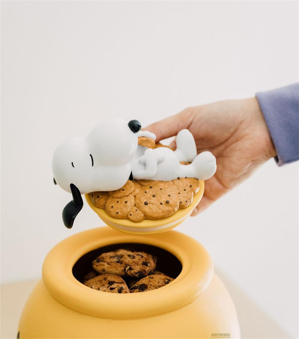 Figura de Snoopy descansa sobre galletas encima de un tarro de galletas amarillo sostenido por una mano.